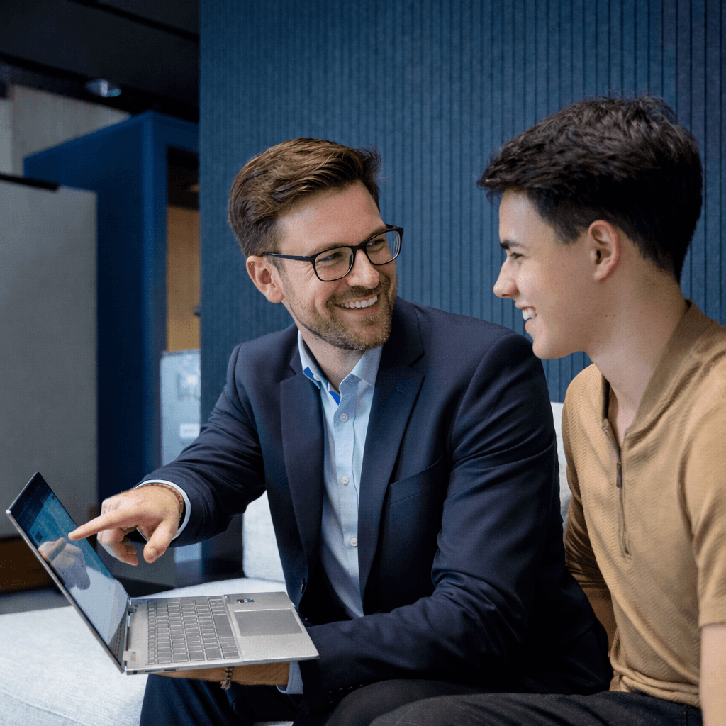 A young person with curly hair smiles at the camera while sitting in front of a laptop against a dark background.