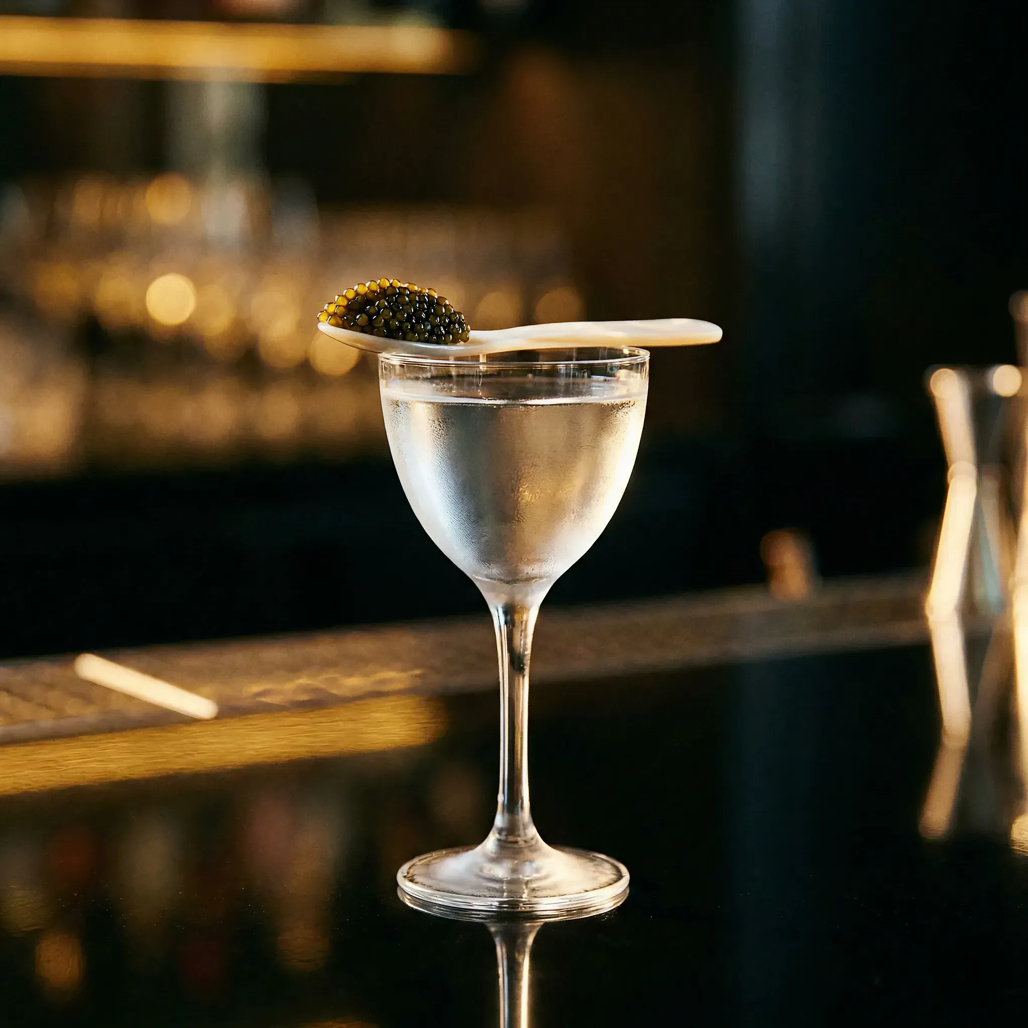 A chilled cocktail glass filled with a clear liquid, topped with a wooden spoon holding dark caviar. Set on a dimly lit bar, exuding elegance and luxury.