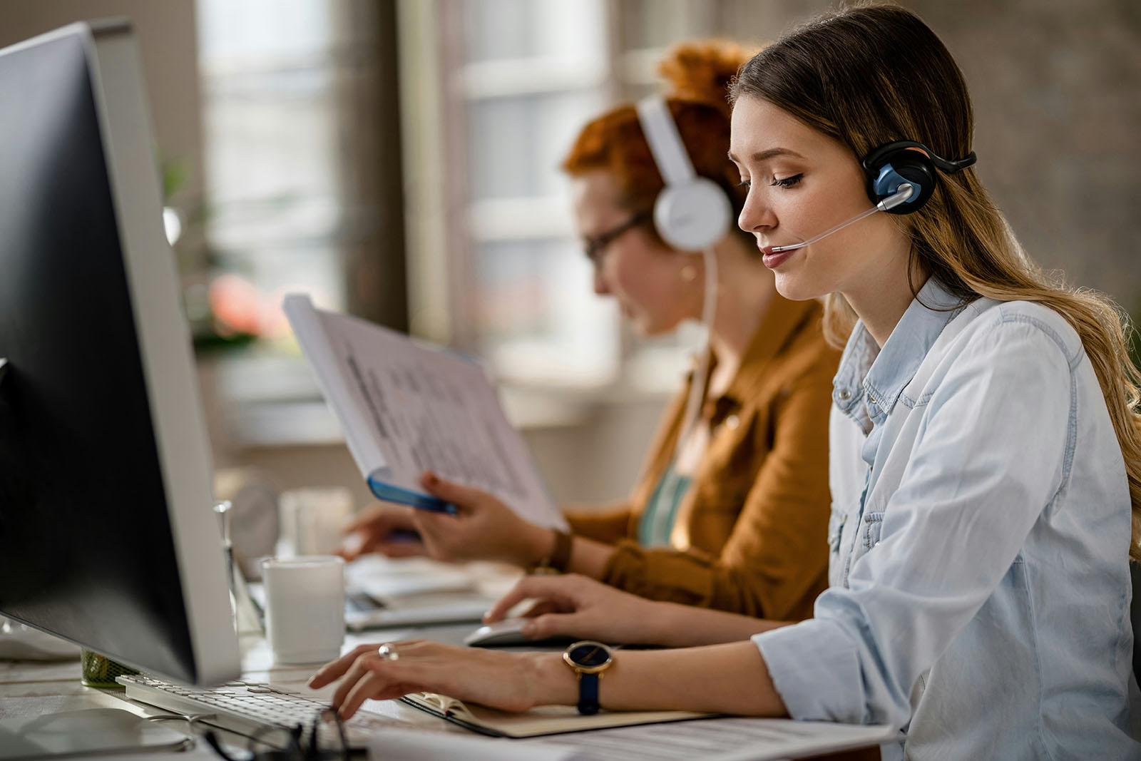 Two women wearing headsets working at computers.