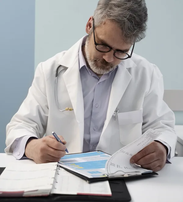 Senior doctor filling in a patient record in a binder at his desk