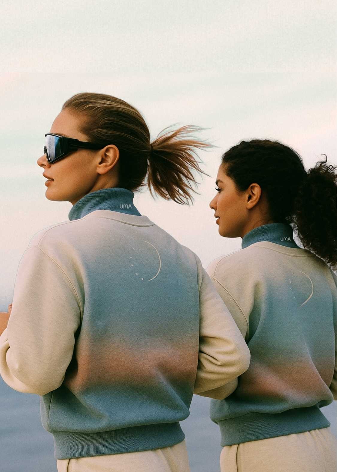 UMA lifestyle photography of two women wearing gradient UMA sweatsuits merch on a beach, representing empowerment, community, and a modern wellness-driven brand aesthetic.