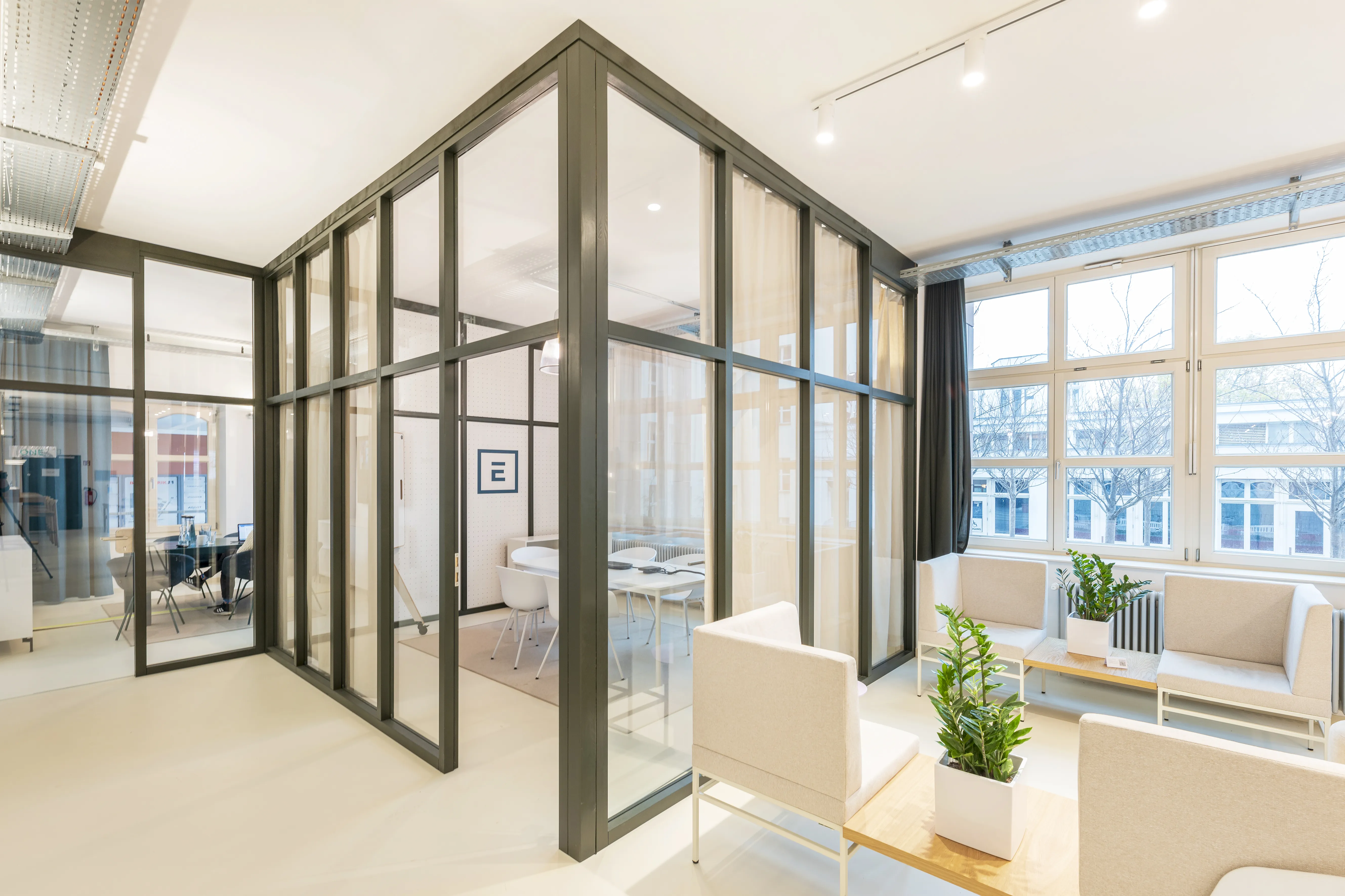 Glass meeting cube at ELEMENT with dark grey steel frames and sheer curtains, next to a lounge with cream armchairs, a sofa bench and a potted plant by large industrial windows.