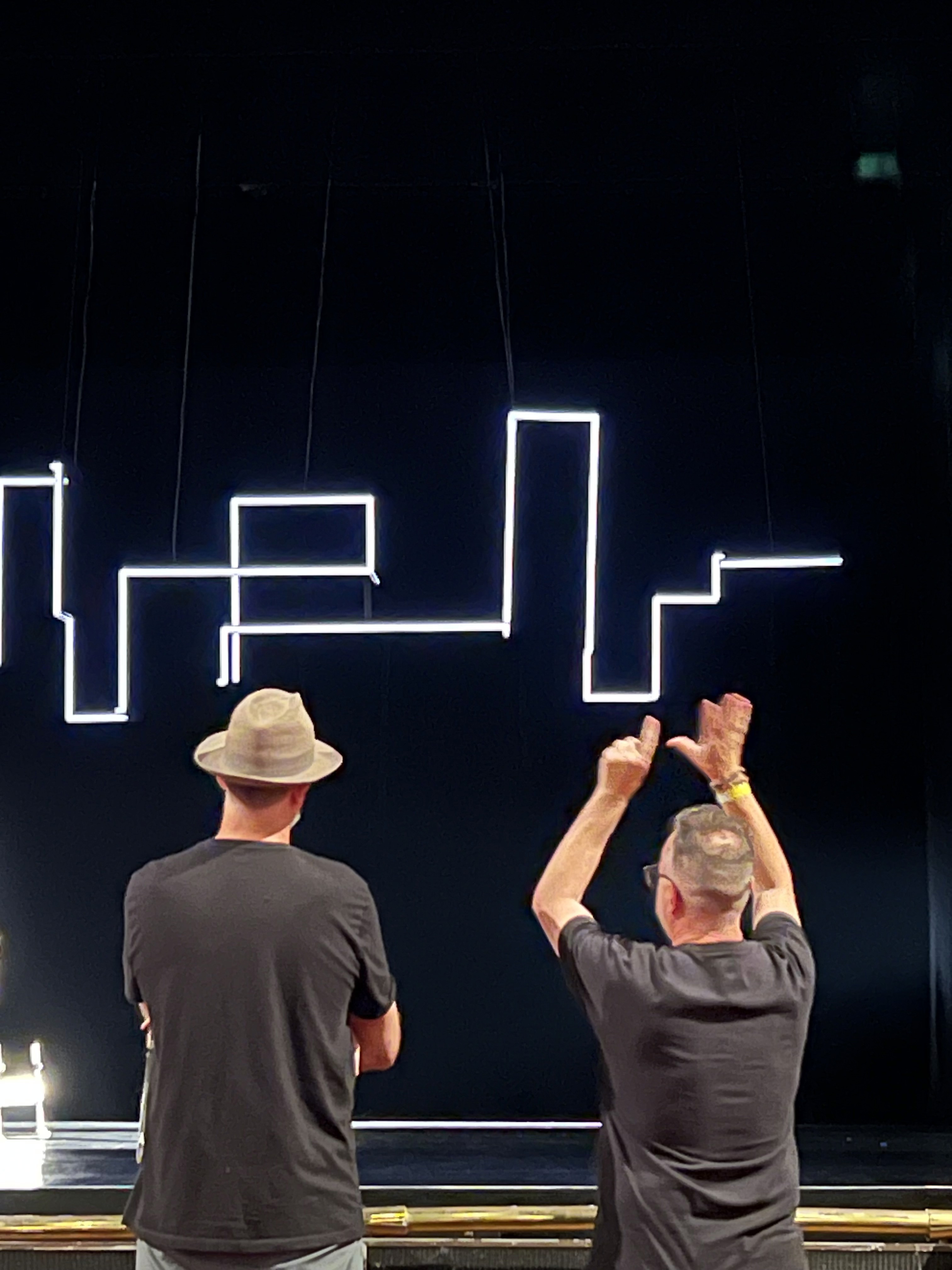 Two people observe a light display on a stage, with visual lines forming abstract shapes in a dark setting.