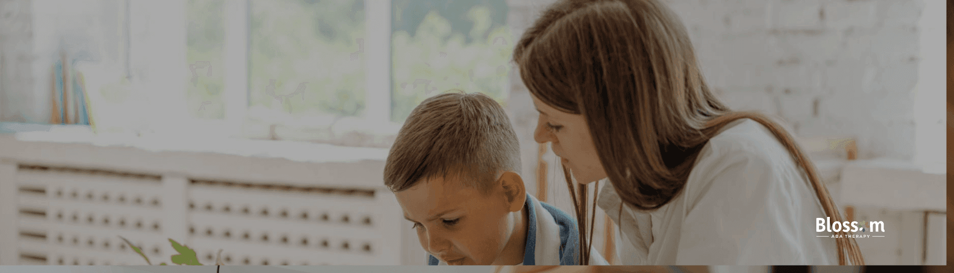Teacher sitting beside a young boy, guiding him through a book at a bright classroom table with school supplies nearby.