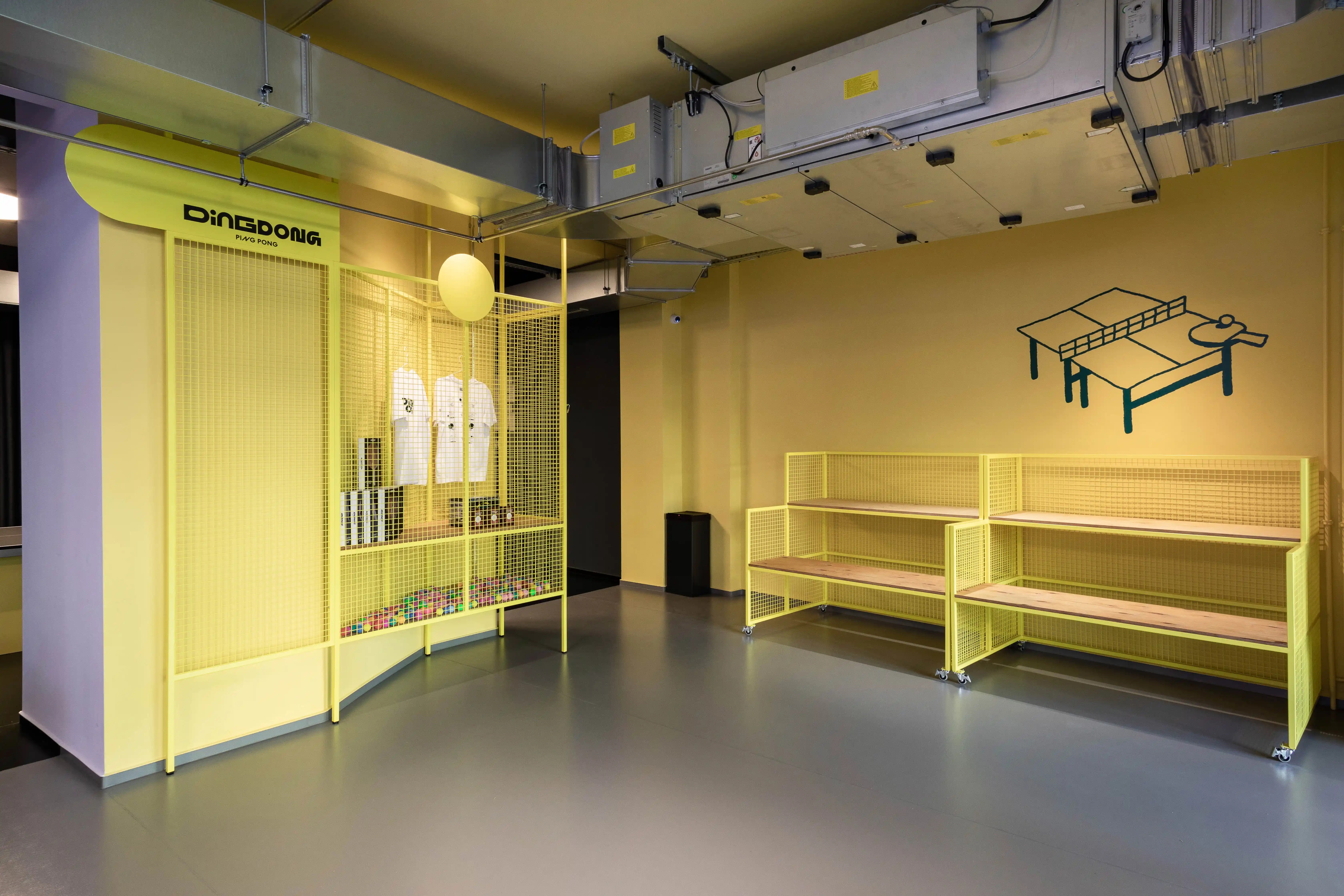 Shop area at DING DONG PING PONG with a yellow mesh kiosk holding shirts and ball tubes, yellow shelf benches and a dark table-tennis-table mural on the yellow wall.