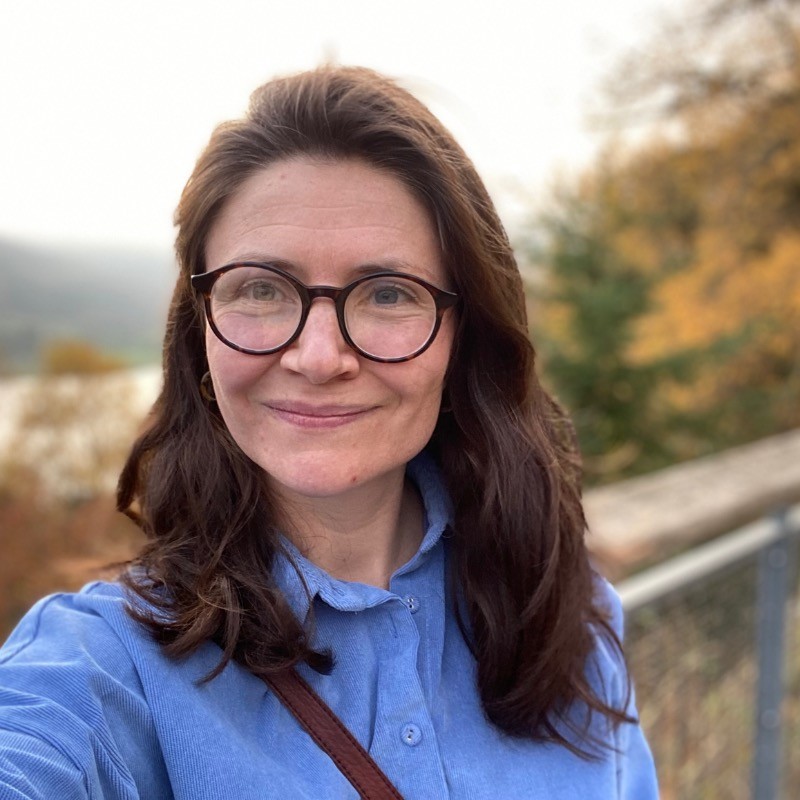Outdoor portrait of a person with shoulder-length dark hair wearing round glasses and a light blue button-up shirt. A brown crossbody strap is visible across the front. The photo is taken on a balcony or raised walkway with a metal railing, with a blurred background of trees showing autumn colours and a distant hillside. Natural daylight and shallow depth of field keep the person in focus while the background is softly blurred.