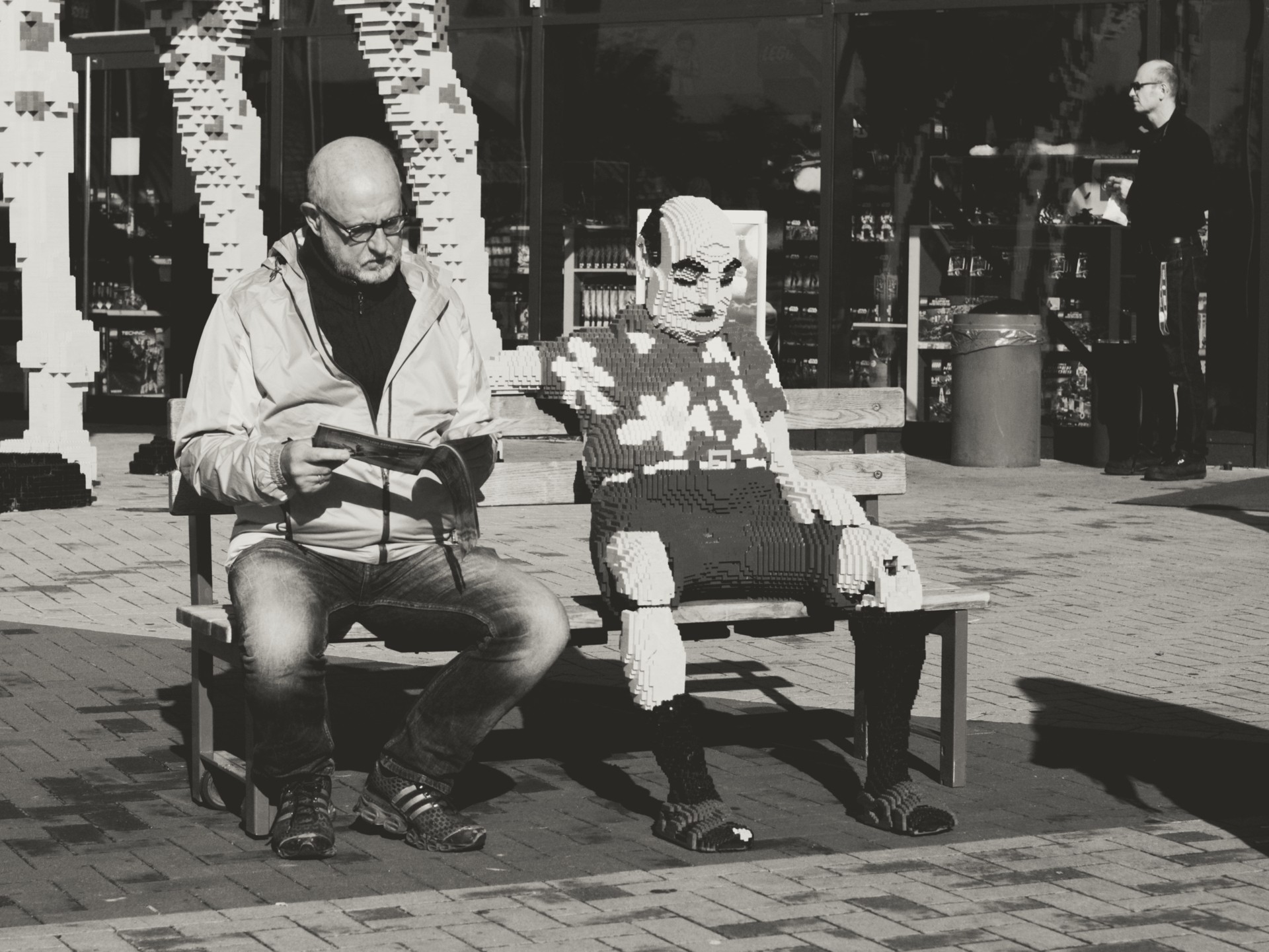 Black-and-white urban photo of a reader on a bench beside a life-sized LEGO sculpture — ideal for content about public art, LEGO installations, street photography, and urban culture.