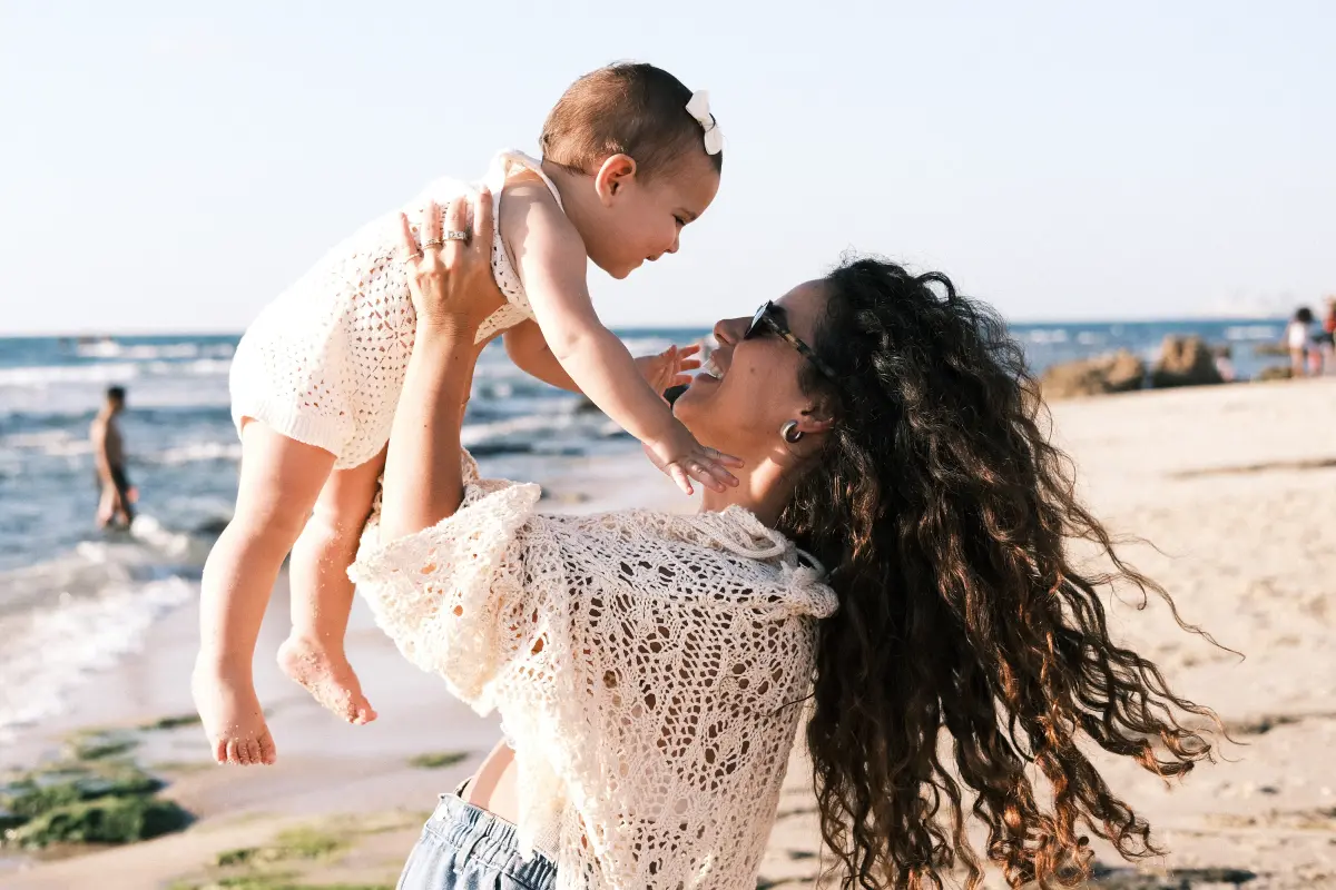 רגע אינטימי ומרגש של אמא המניפה את תינוקה באוויר על חוף הים בשעת בין ערביים. התמונה מאופיינת בתאורה טבעית רכה ובאווירת לייף-סטייל נינוחה על רקע החול והגלים, מאת Shiriz Photography הצלמת שירי עציץ.