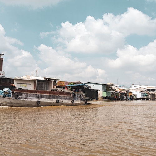 Pemandangan tepi sungai dengan perahu dan bangunan di atas pancang di samping air di bawah langit yang sebagian berawan.