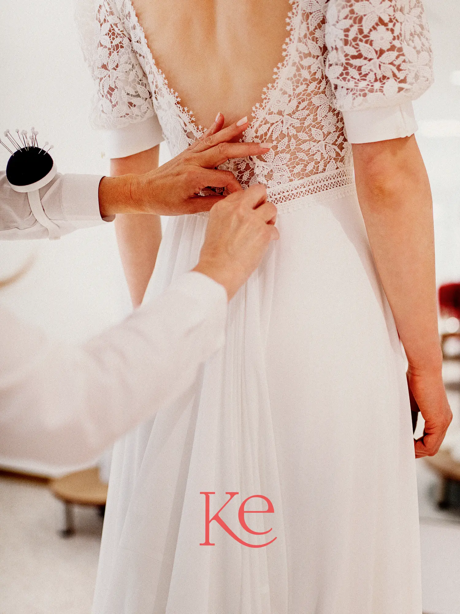 Detailaufnahme einer Brautkleid-Anprobe bei Kerst Hochzeitsmode: Eine Schneiderin justiert das filigrane Spitzenoberteil eines weißen Hochzeitskleides, ergänzt durch das rote Ke-Logo.