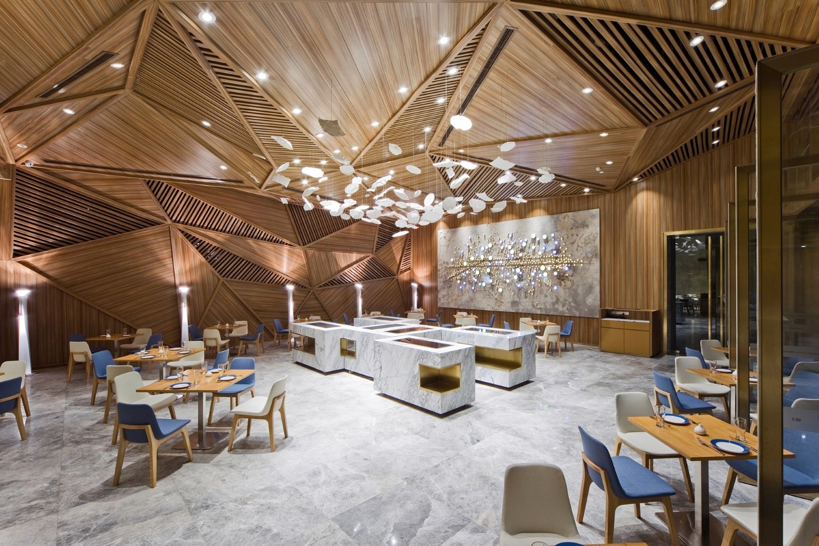 A modern restaurant interior featuring a geometric wooden ceiling, elegant marble tables, contemporary blue and beige chairs, and a striking wall installation with artistic lighting, creating a luxurious dining atmosphere.