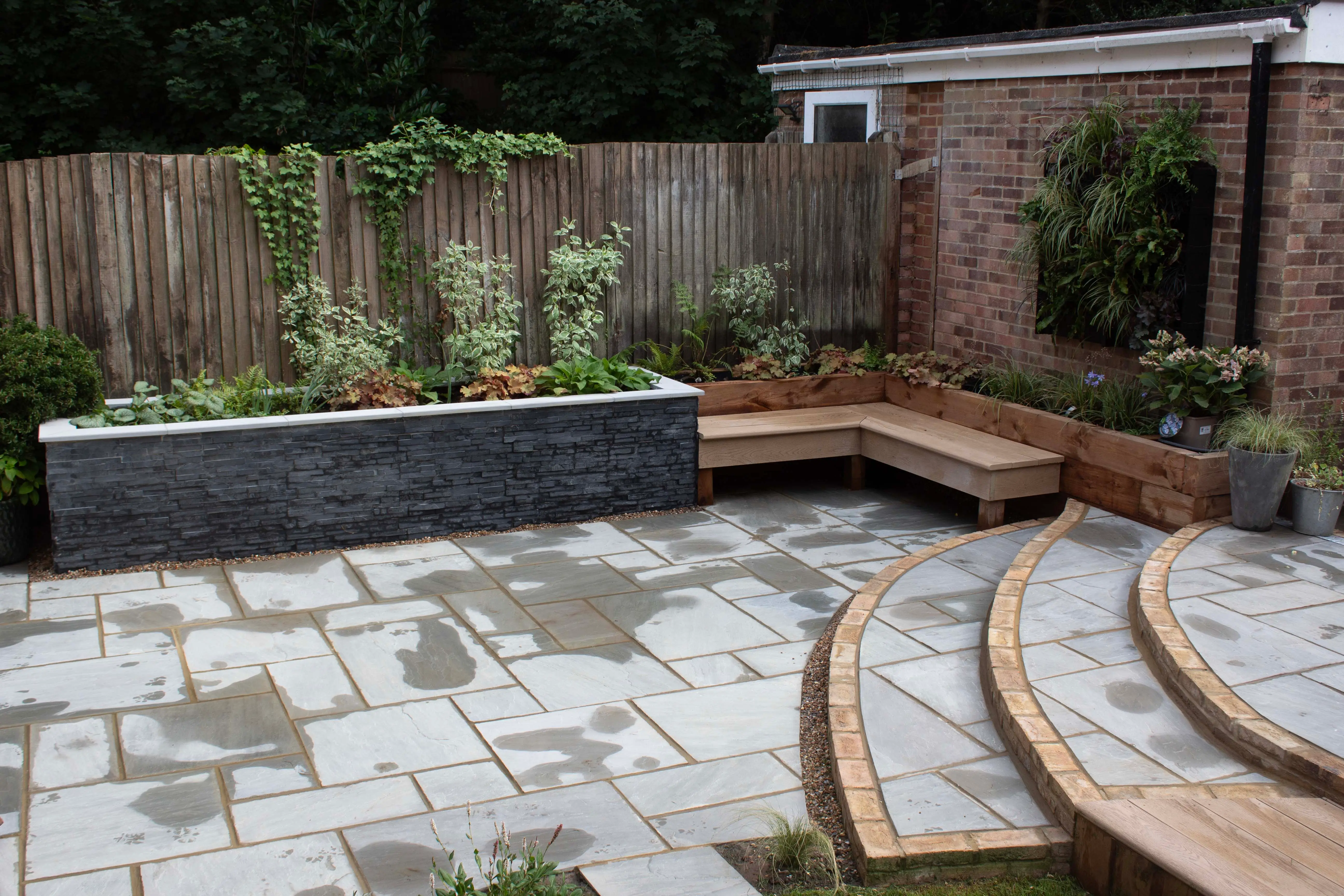 A patio area features stone flooring, a curved wooden bench, and plants in a garden setting with a wooden fence.