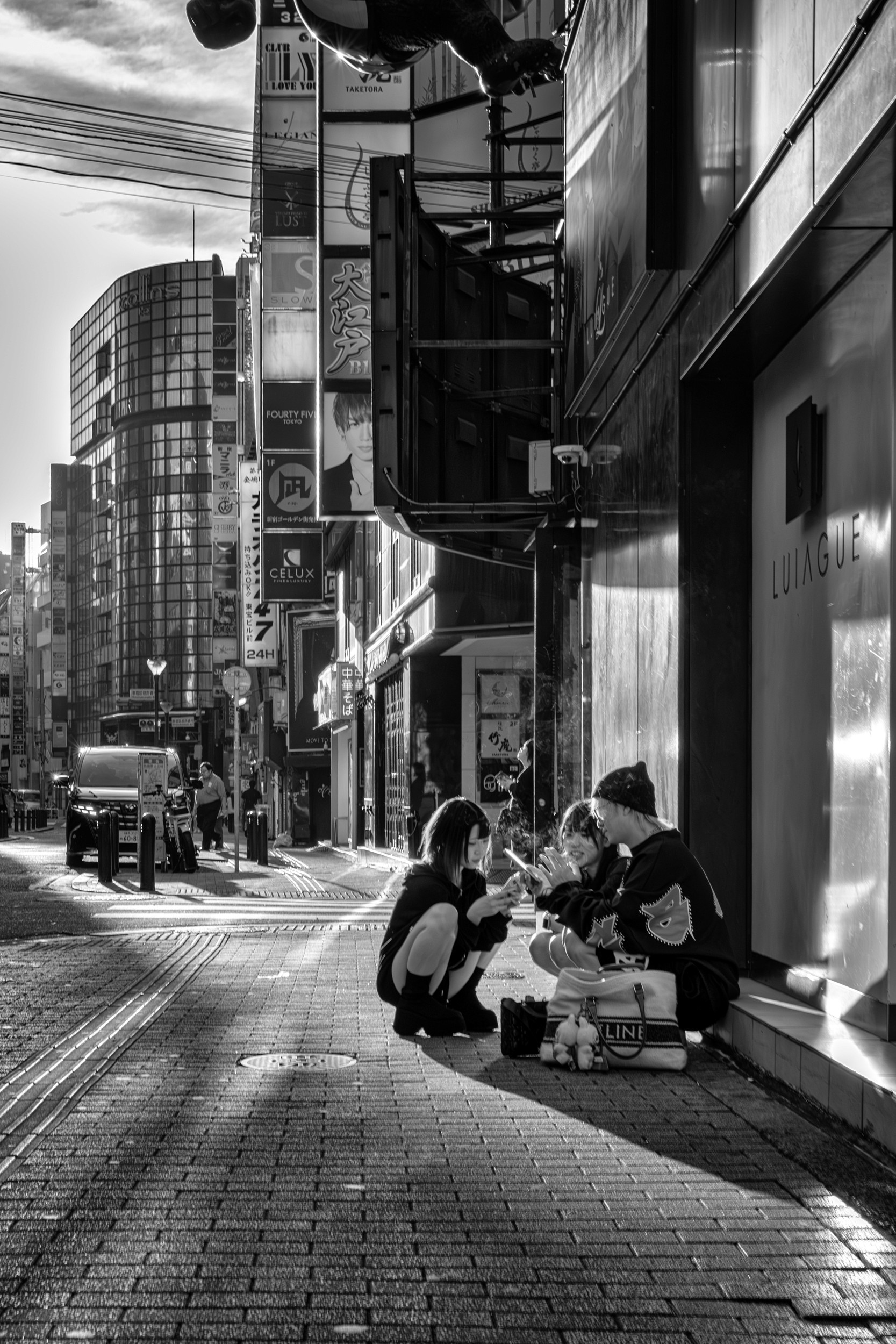 Black and white street photograph of three young people crouching on a curb looking at a phone.