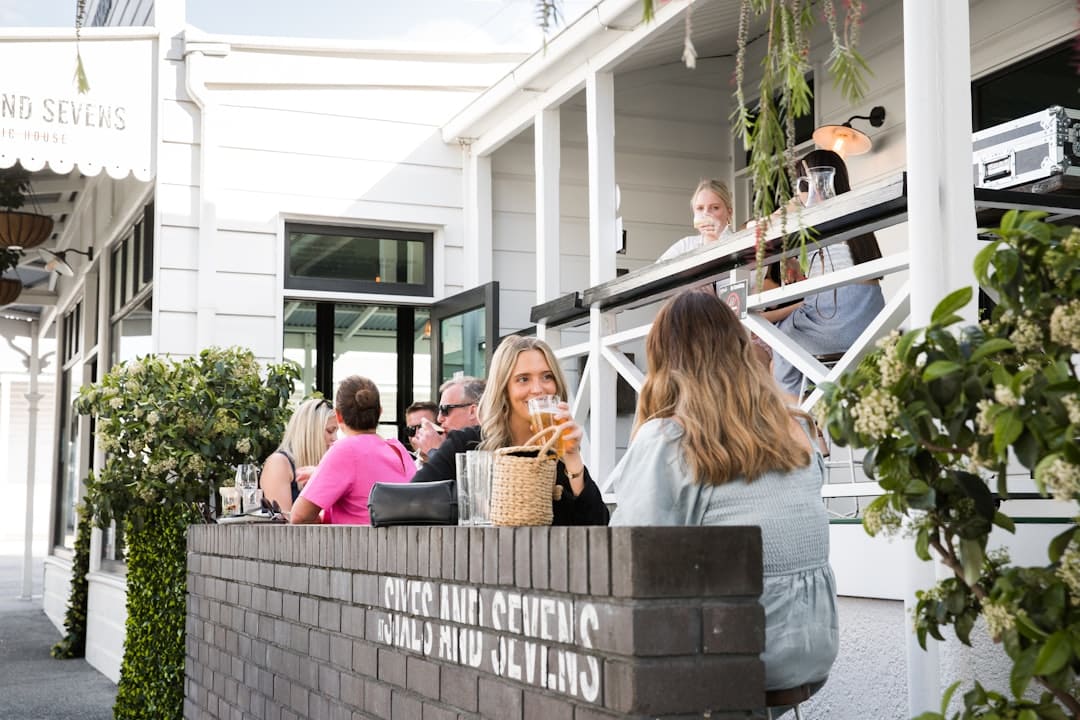 People enjoying drinks and socializing at an outdoor seating area of a white modern café or bar named 'Sixes and Sevens'.