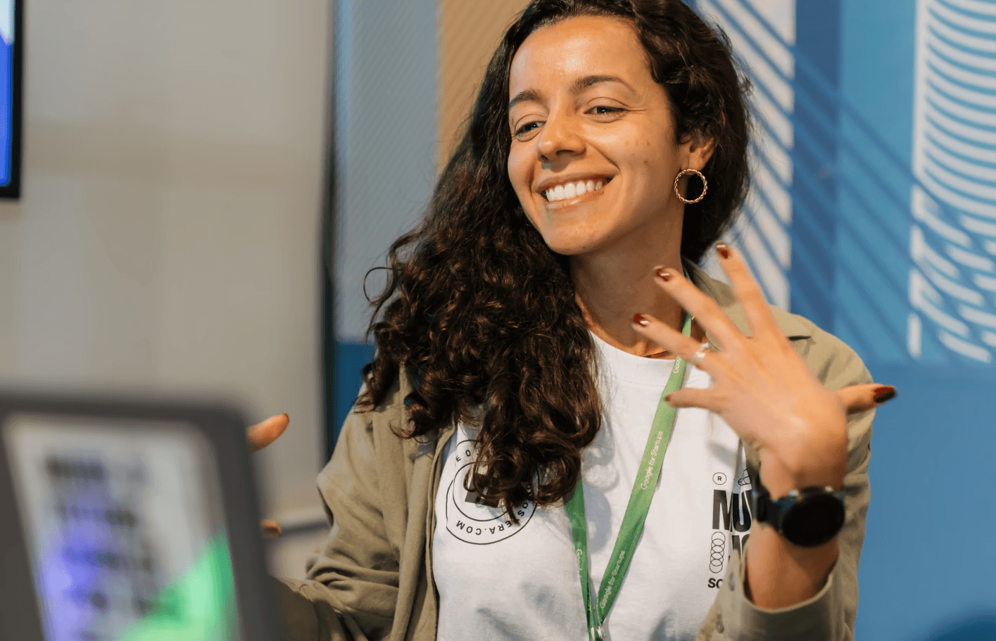 Uma pessoa sorridente com cabelos longos está sentada em uma cadeira, cercada por plantas e com uma vista da cidade ao fundo.