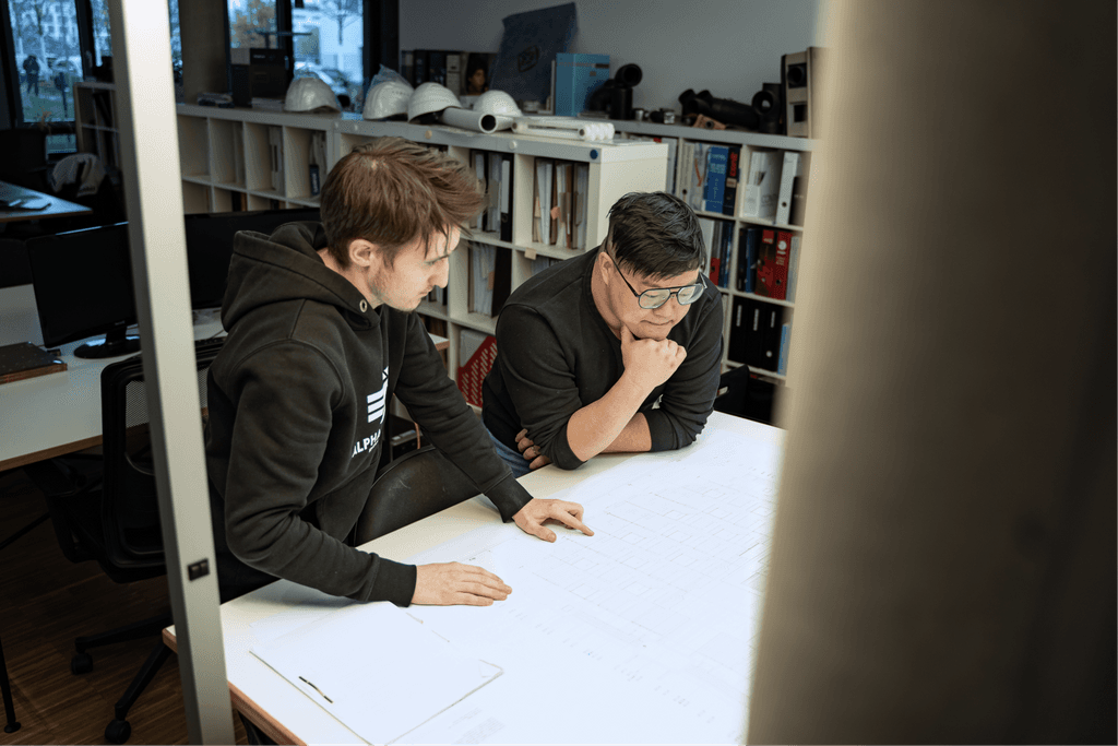 Zwei Mitarbeitende der kba Architekten und Ingenieure GmbH beugen sich über großformatige technische Baupläne und analysieren Details; im Hintergrund stehen Regale mit Ordnern und Sicherheitshelmen in einem modernen Büro.