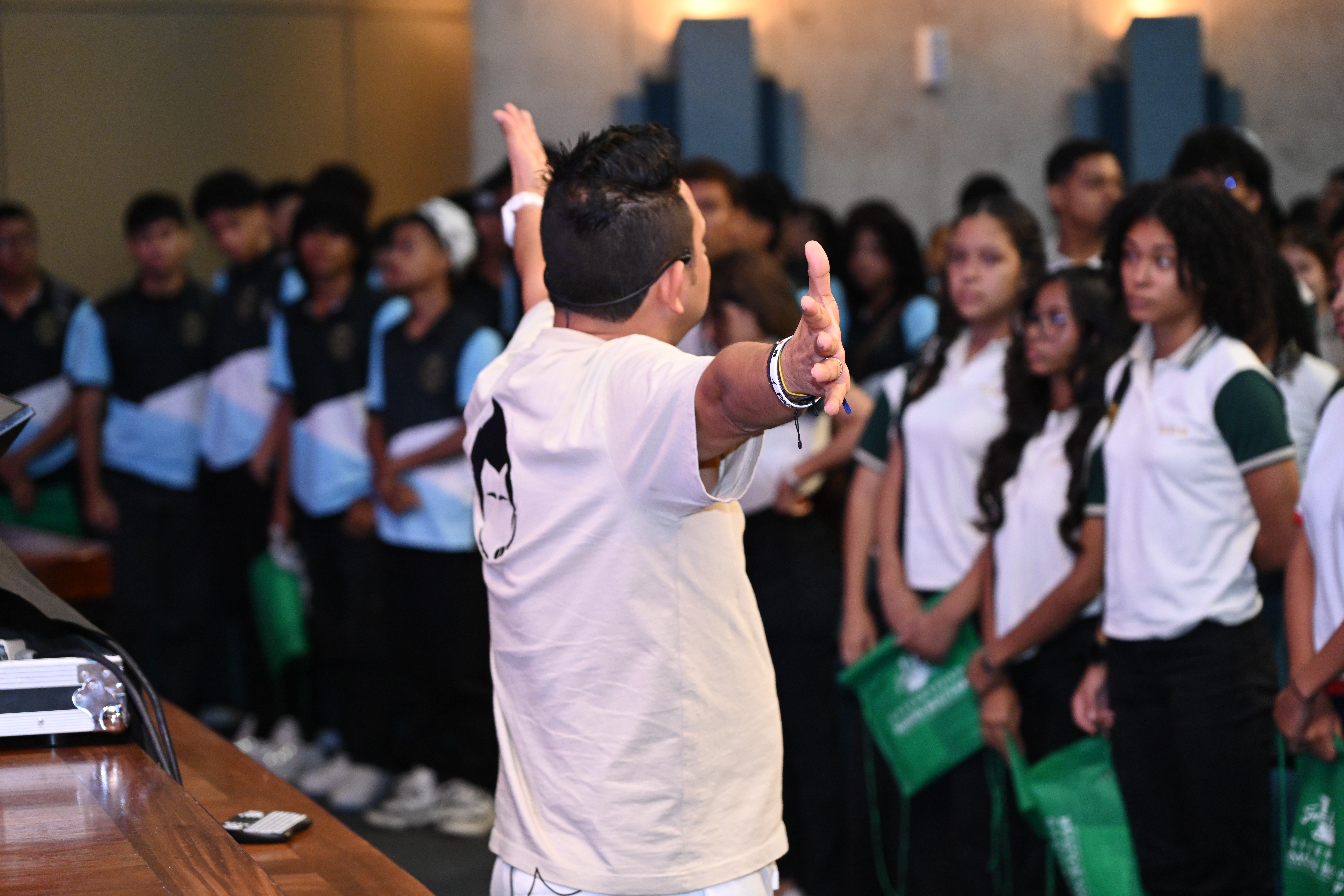 Jorge Baena de espaldas, con los brazos extendidos, hablando con energía frente a un grupo de estudiantes uniformados que lo observan atentamente durante una conferencia motivacional.