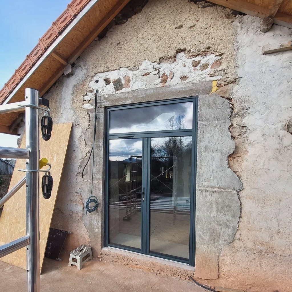 Façade en rénovation avec porte-fenêtre noire à deux vantaux et partie haute fixe, entourée d’un mur partiellement enduit.