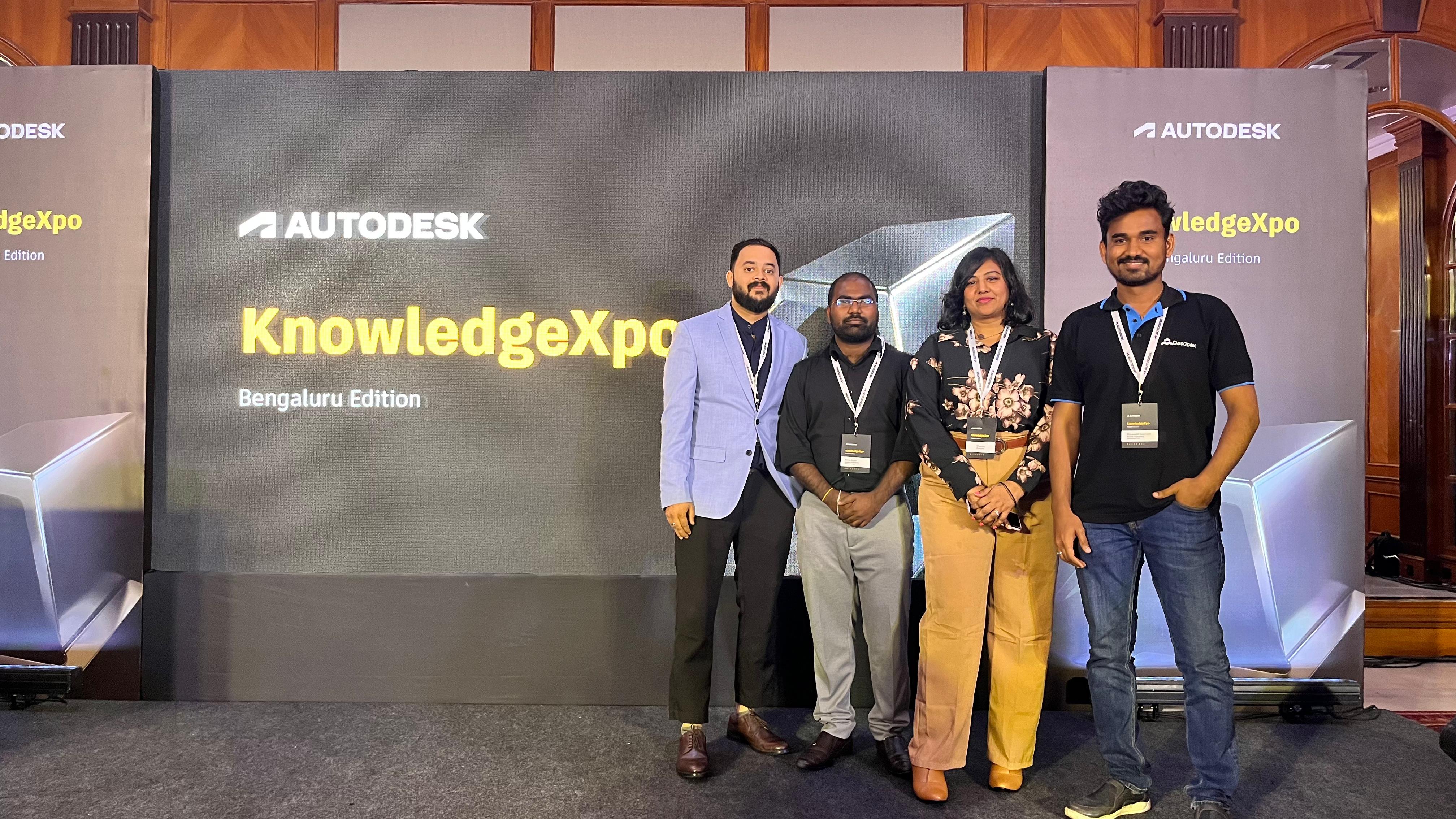 A man stands while two people sit at a table in an office setting, smiling and engaged in conversation. The mood is friendly and collaborative.