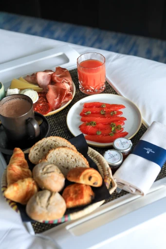 Un plateau de petits-déjeuners présente des tranches de saumon fumé garnies de câpres, accompagnées de charcuterie, de pains assortis, de viennoiseries et d’un verre de jus d’orange, mettant en avant un cadre de service en chambre de luxe dans un hôtel.