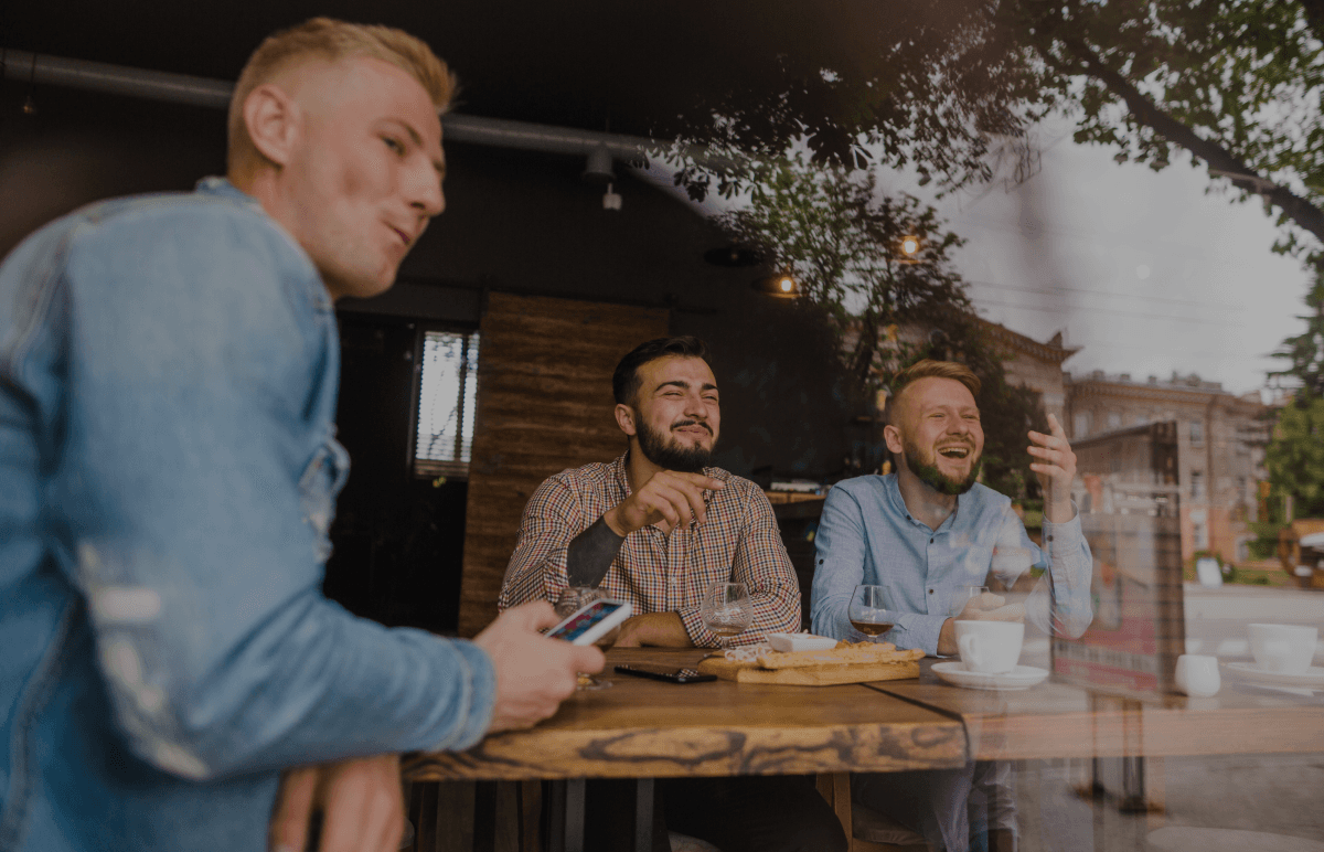 Una escena informal de tres amigos riendo y hablando mientras están sentados en una mesa de café. La foto fue tomada desde fuera, mirando a través de la ventana, capturando una atmósfera cálida y relajada. Una persona sostiene un teléfono, y hay comida y bebidas en la mesa.