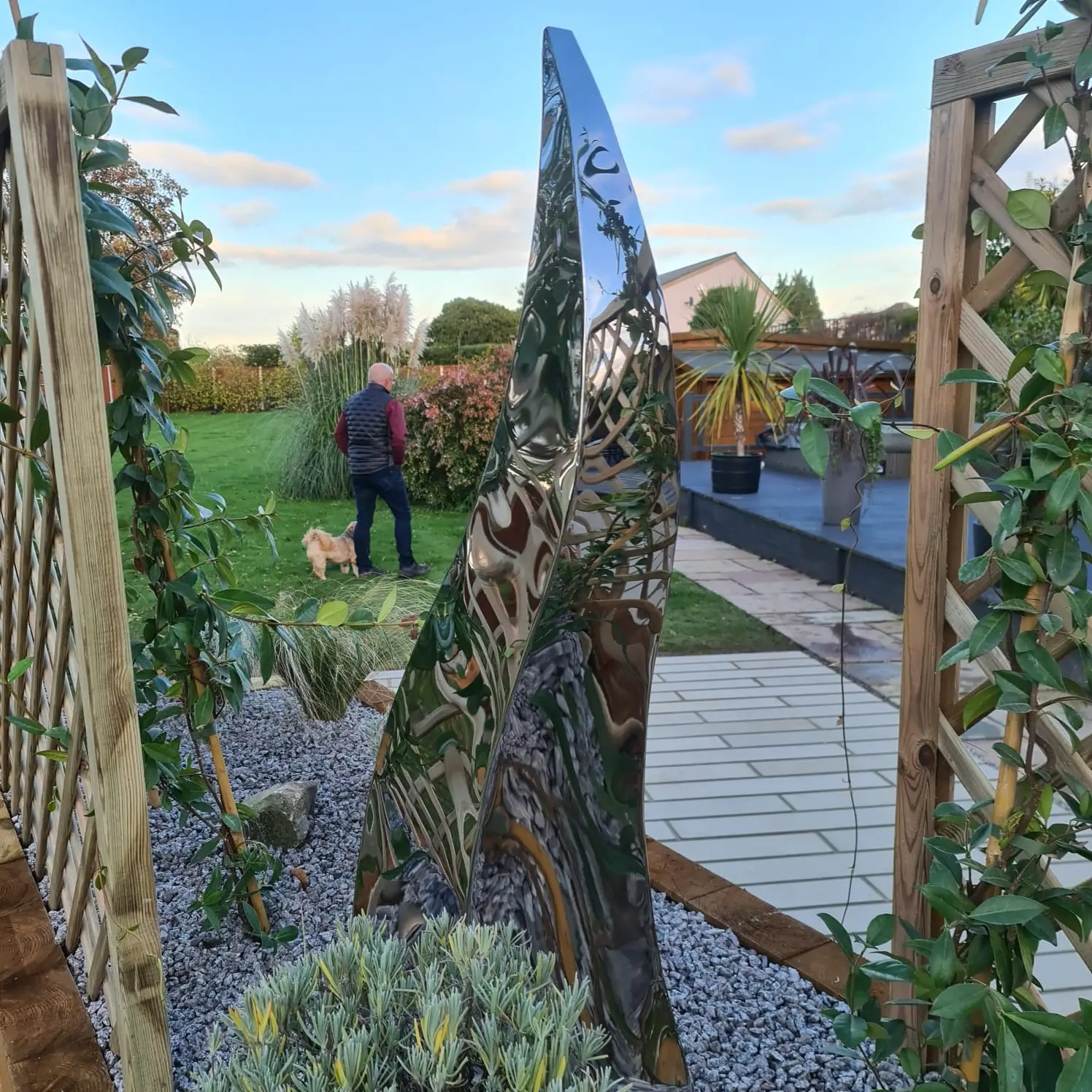 A person stands near a large, artistic structure surrounded by plants and greenery in an outdoor setting.