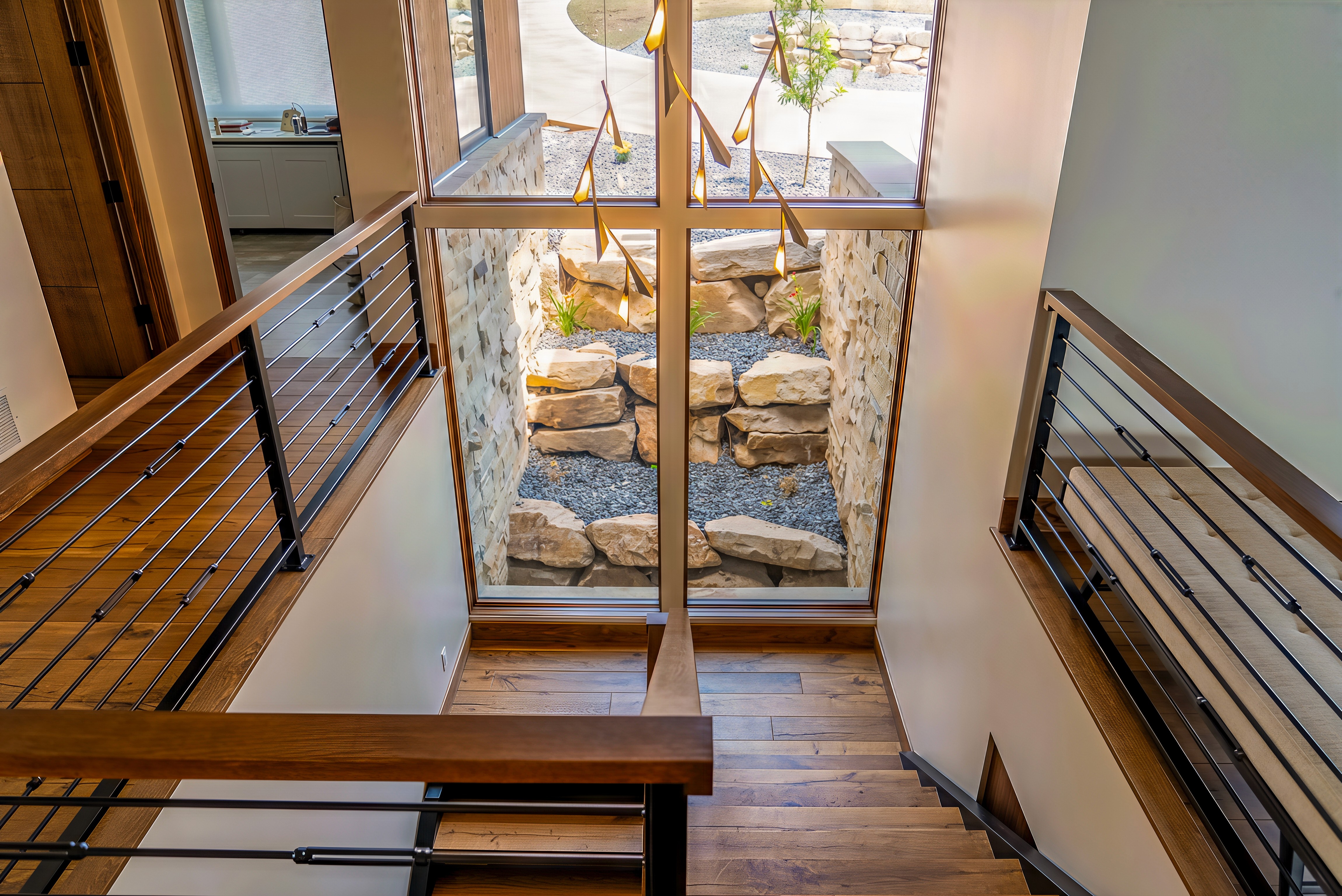 Modern luxury home interior featuring a multi-level staircase with warm wood floors, metal railing accents, and a large floor-to-ceiling window overlooking a stone-lined courtyard, illuminated by a sculptural pendant light.
