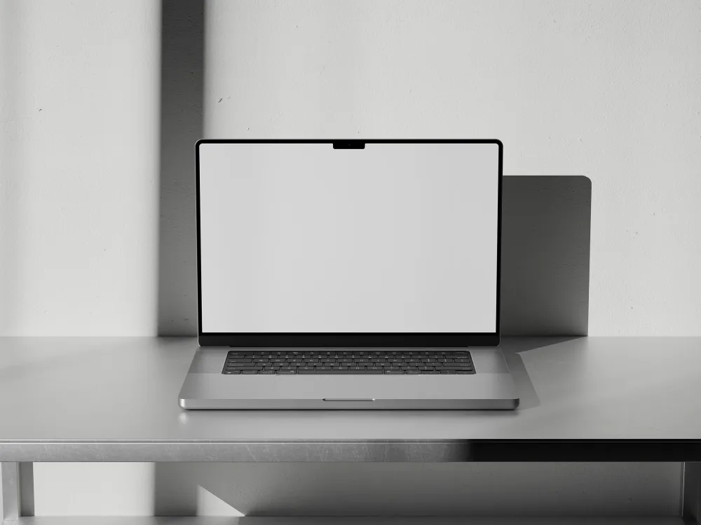 MacBook Pro on a metal desk, displaying a minimal website design with beige background, bold typography, and black-and-white images. Soft shadows cast on a textured white wall.