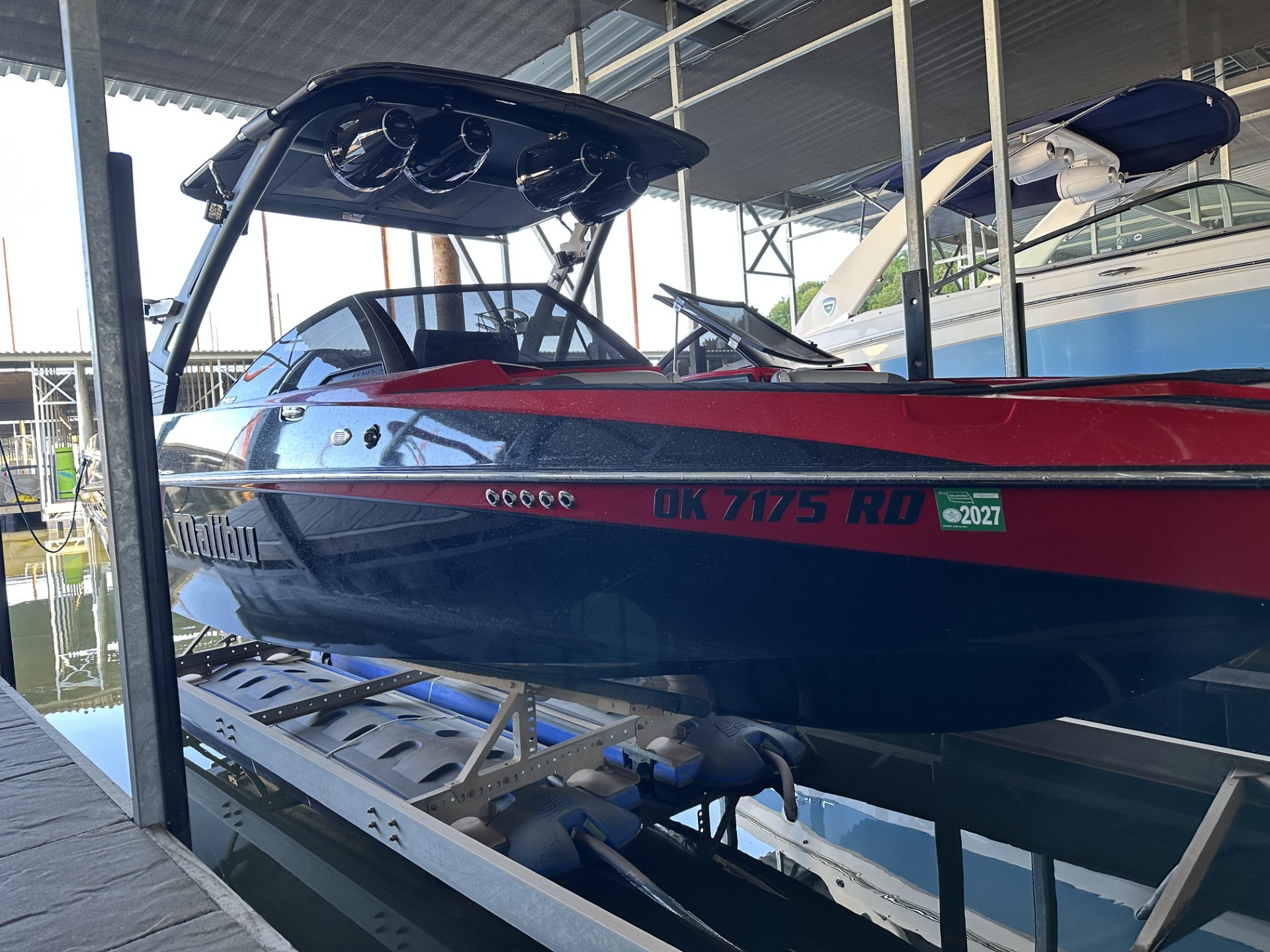 A sleek red and blue speedboat with a shiny black canopy is docked in a marina, supported by a metal lift system, with water reflecting beneath.