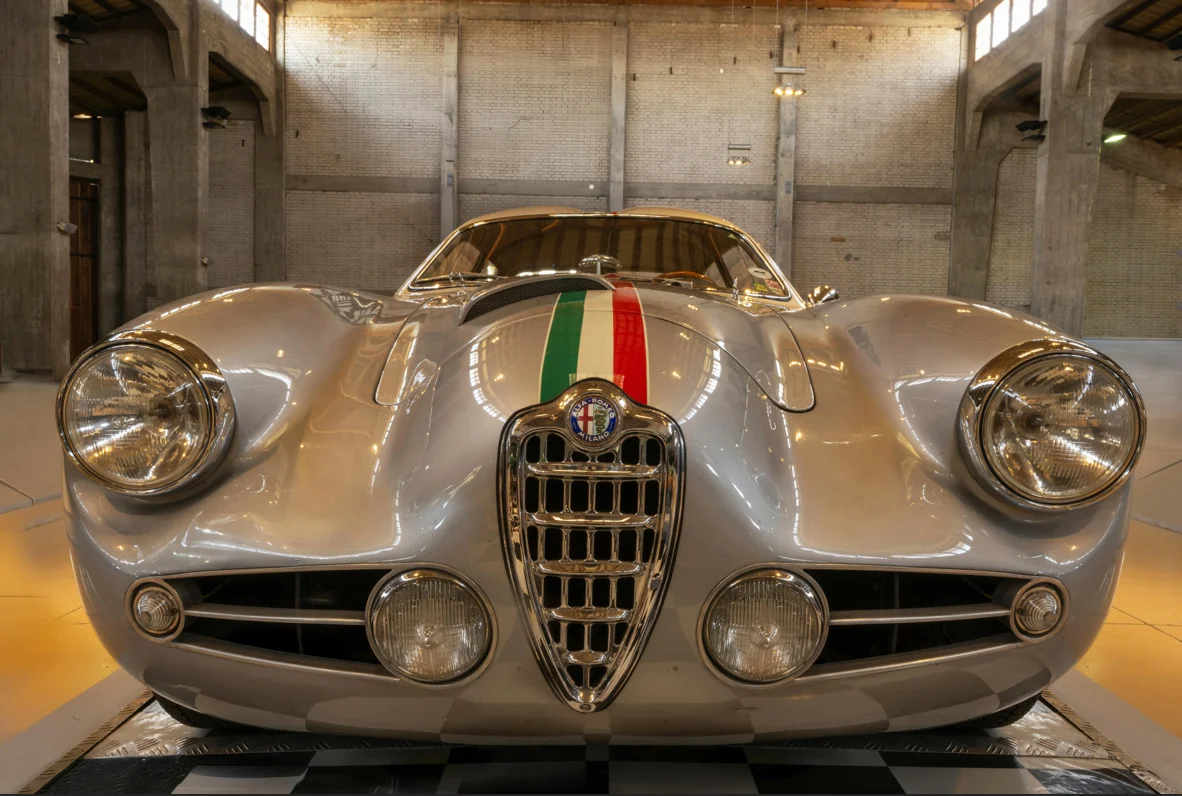 Une Alfa Romeo classique exposée dans un vaste espace industriel. Sa face avant sculptée, marquée par la calandre emblématique et une bande tricolore, met en valeur le charme intemporel du design italien et l’héritage sportif de la marque.