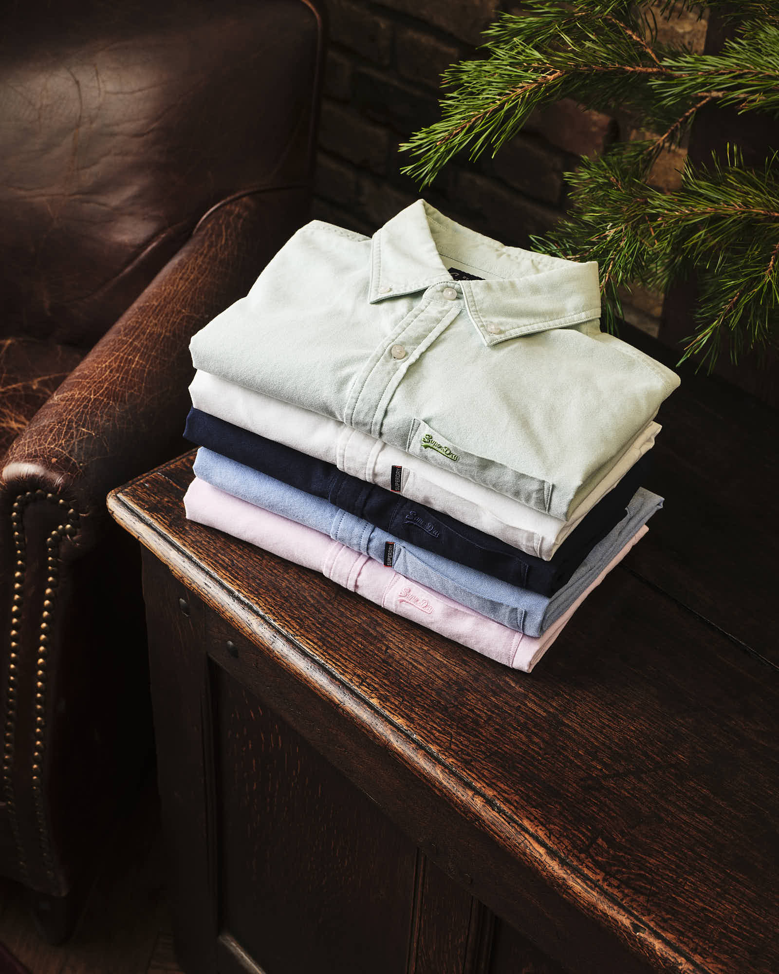 Folded shirts on rustic wooden table with leather chair and pine branches.