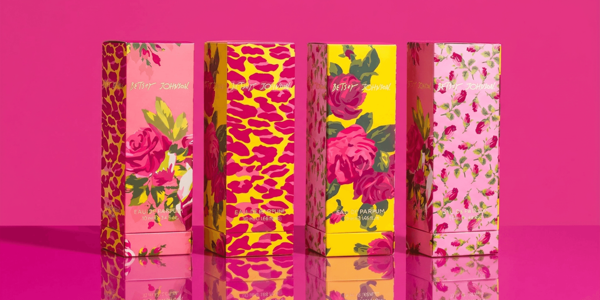Markenwelt Betsey Johnson Parfum – vier Eau-de-Parfum-Faltschachteln in Reihe vor pinkfarbenem Hintergrund, mit gemalten Rosen-Illustrationen, Rosenknospen-Mustern und Leoparden-Prints in Pink, Gelb und Magenta, jeweils mit handschriftlichem „Betsey Johnson"-Logo.