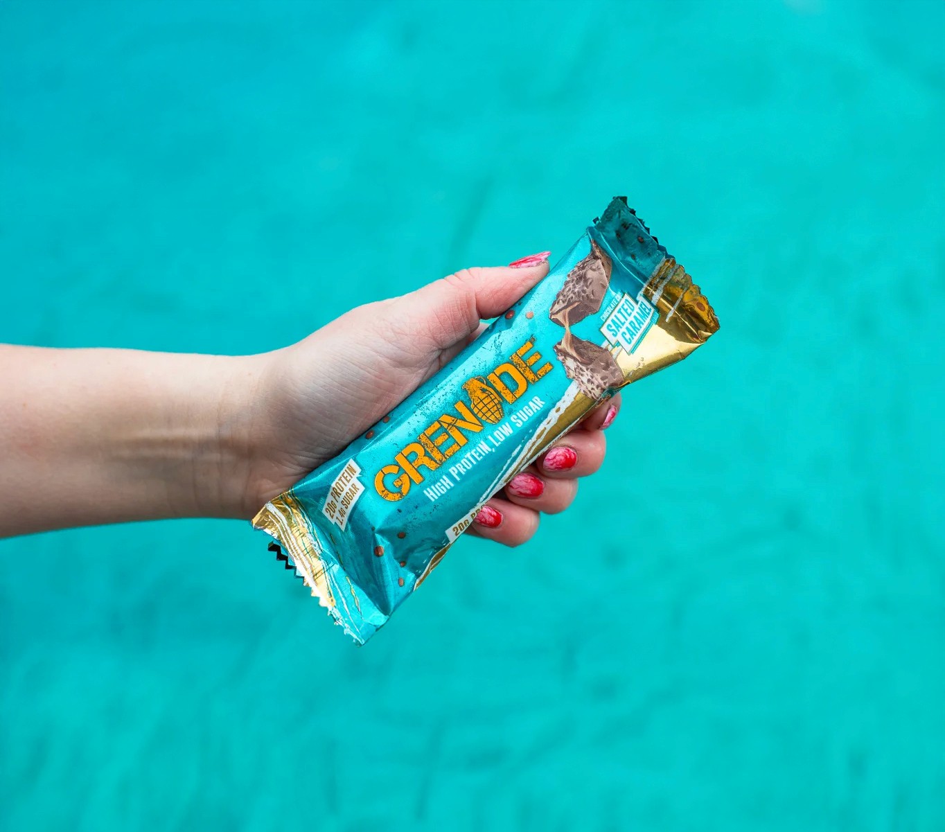 A hand with red-tipped fingernails holds a turquoise and gold "Grenade" high protein bar horizontally against a vibrant, textured teal background. The wrapper for the "Chocolate Chip Salted Caramel" flavor clearly displays text highlighting "20g Protein" and "Low Sugar."