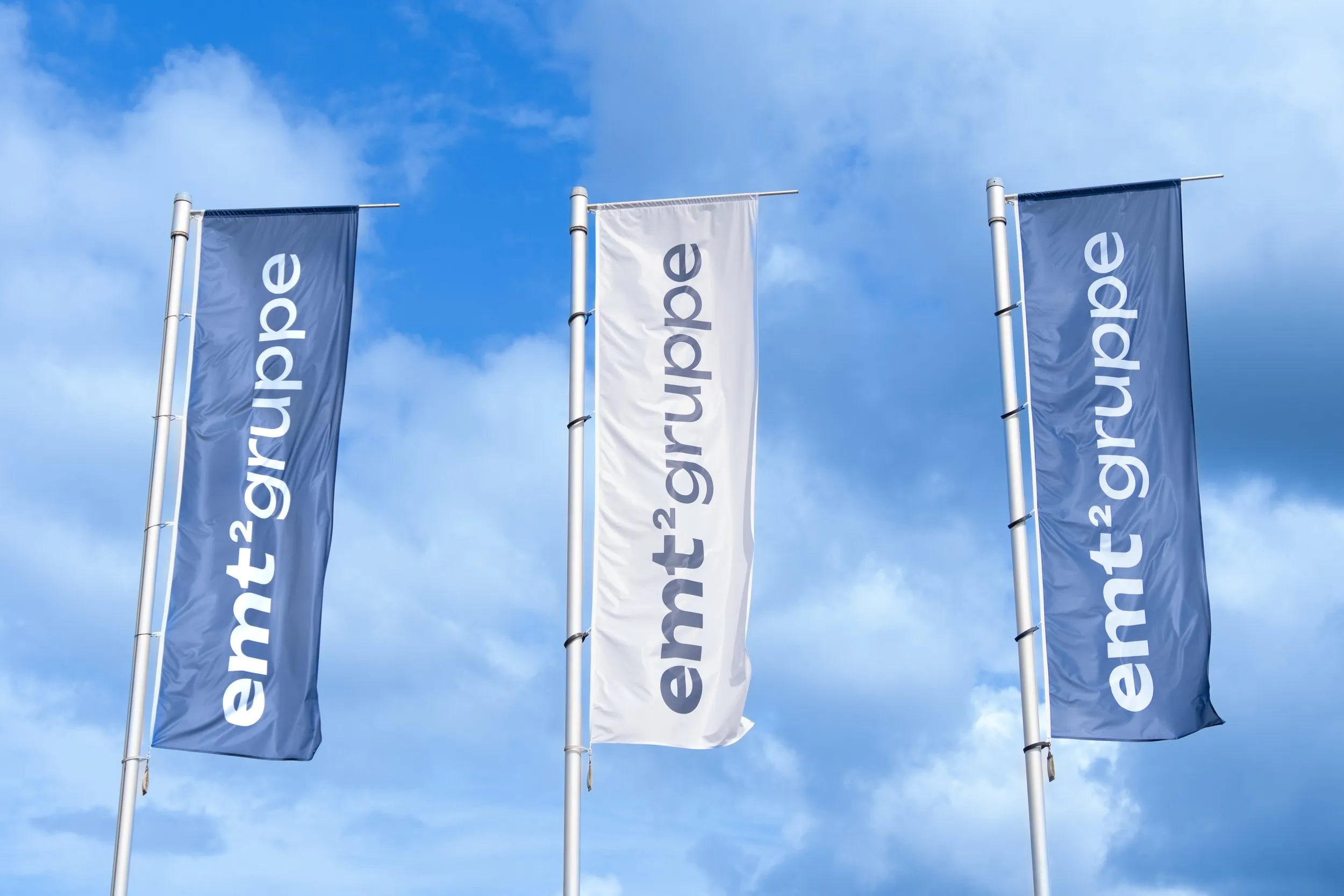 Drei Fahnen mit dem Logo "emt²gruppe" vor blauem Himmel