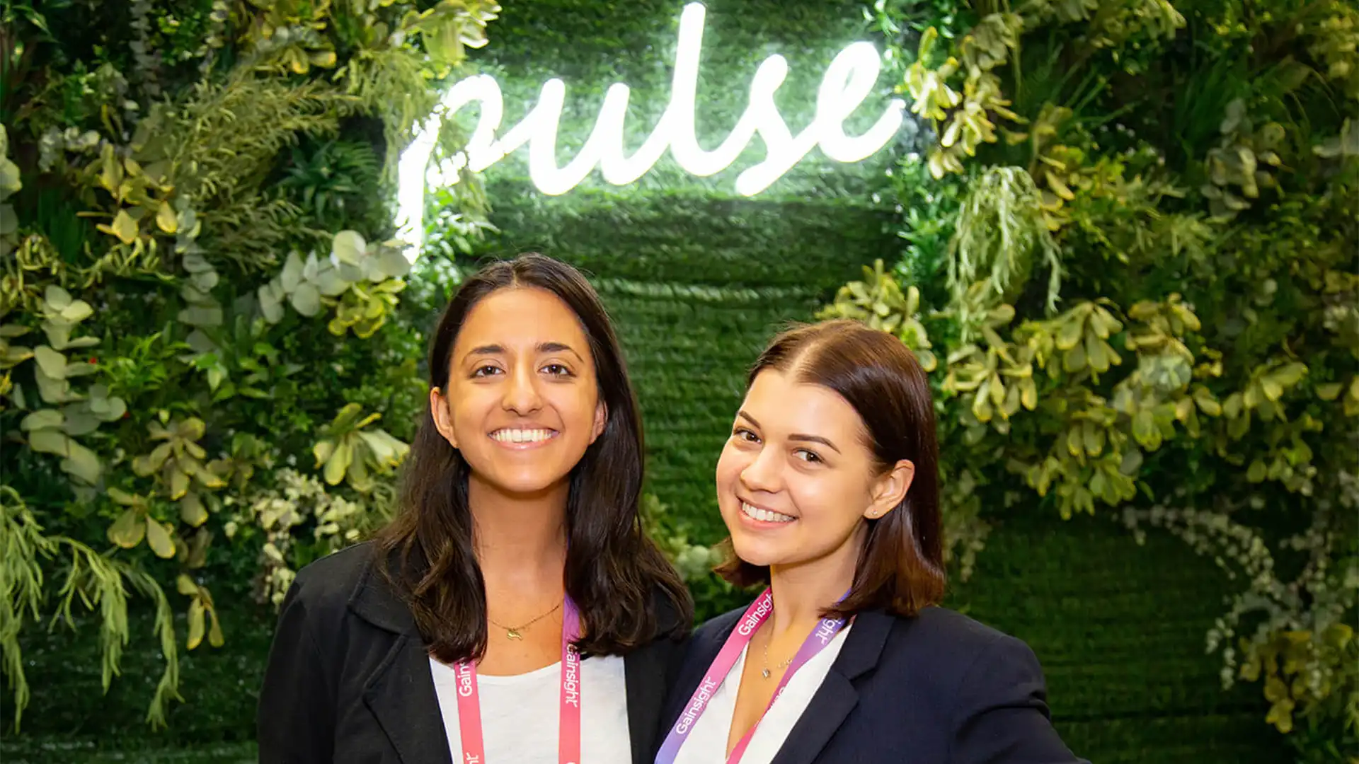 Two conference attendees posing in front of a green Gainsight Pulse feature wall.