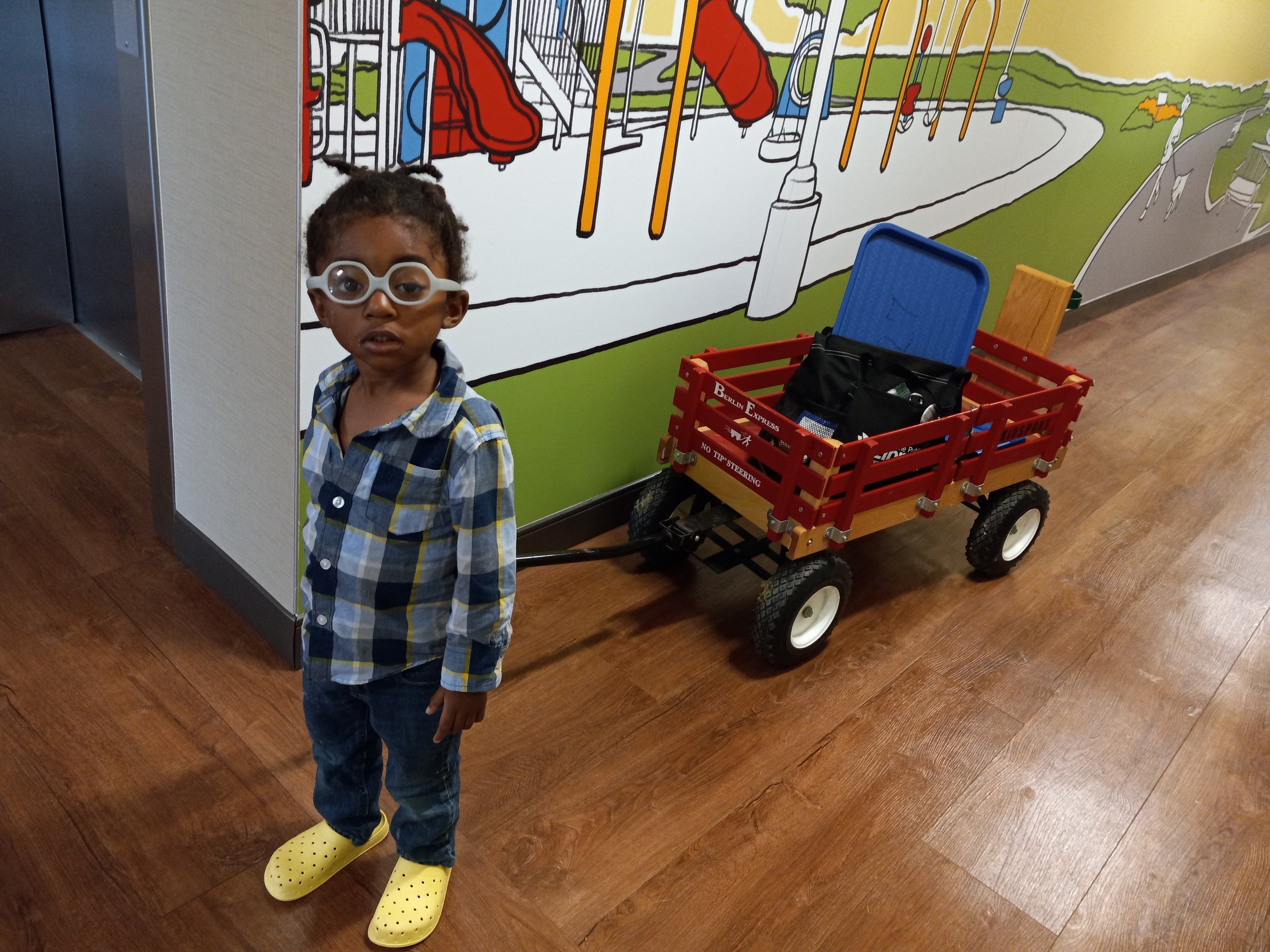 Alt tag: Young child wearing glasses stands indoors near a colorful playground mural, with a red pull wagon carrying personal items behind them on a wooden floor.