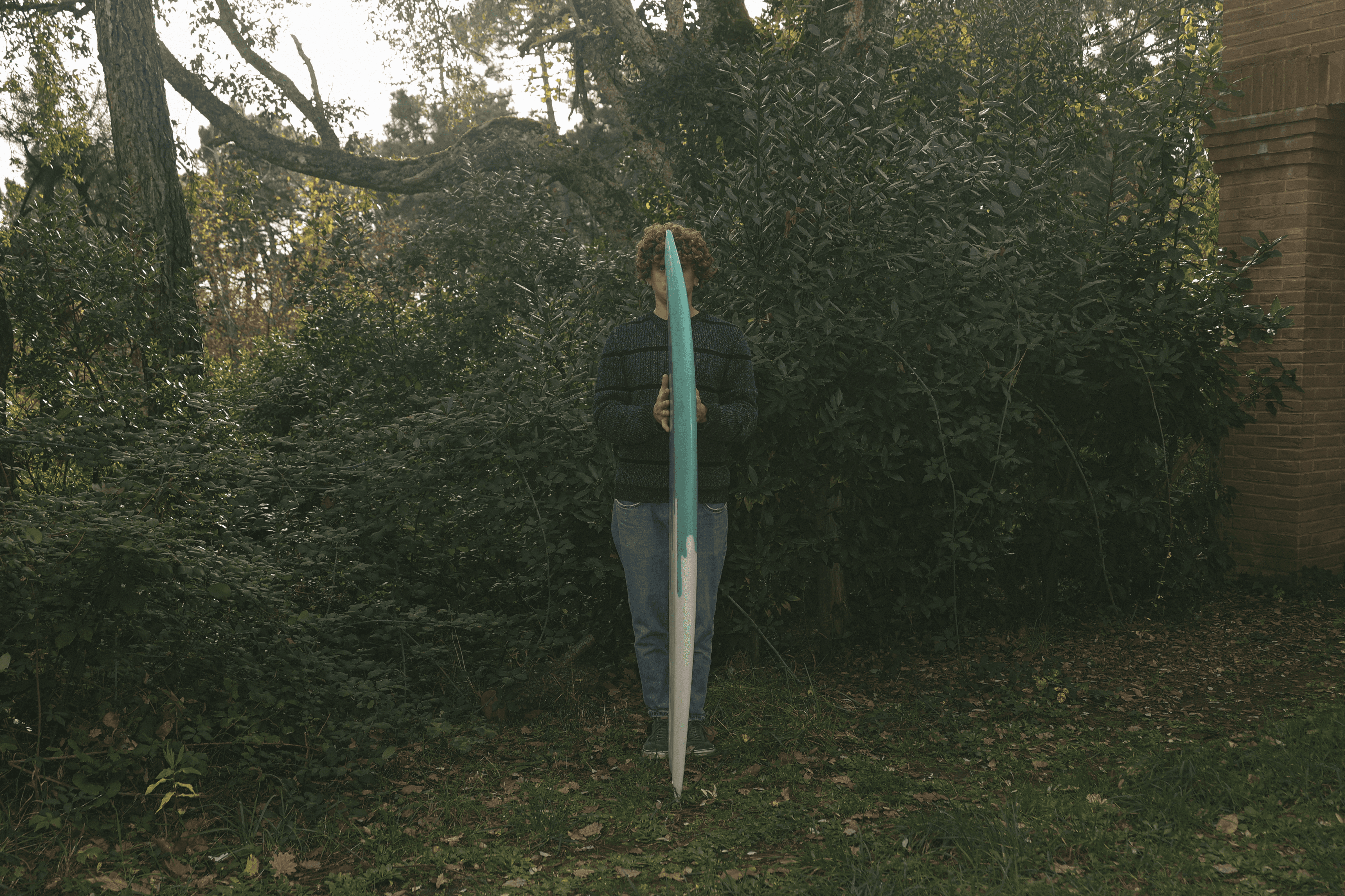 Una persona in piedi in un ambiente boschivo tiene verticalmente una tavola da surf verde acqua e bianca, coprendo parte del viso.