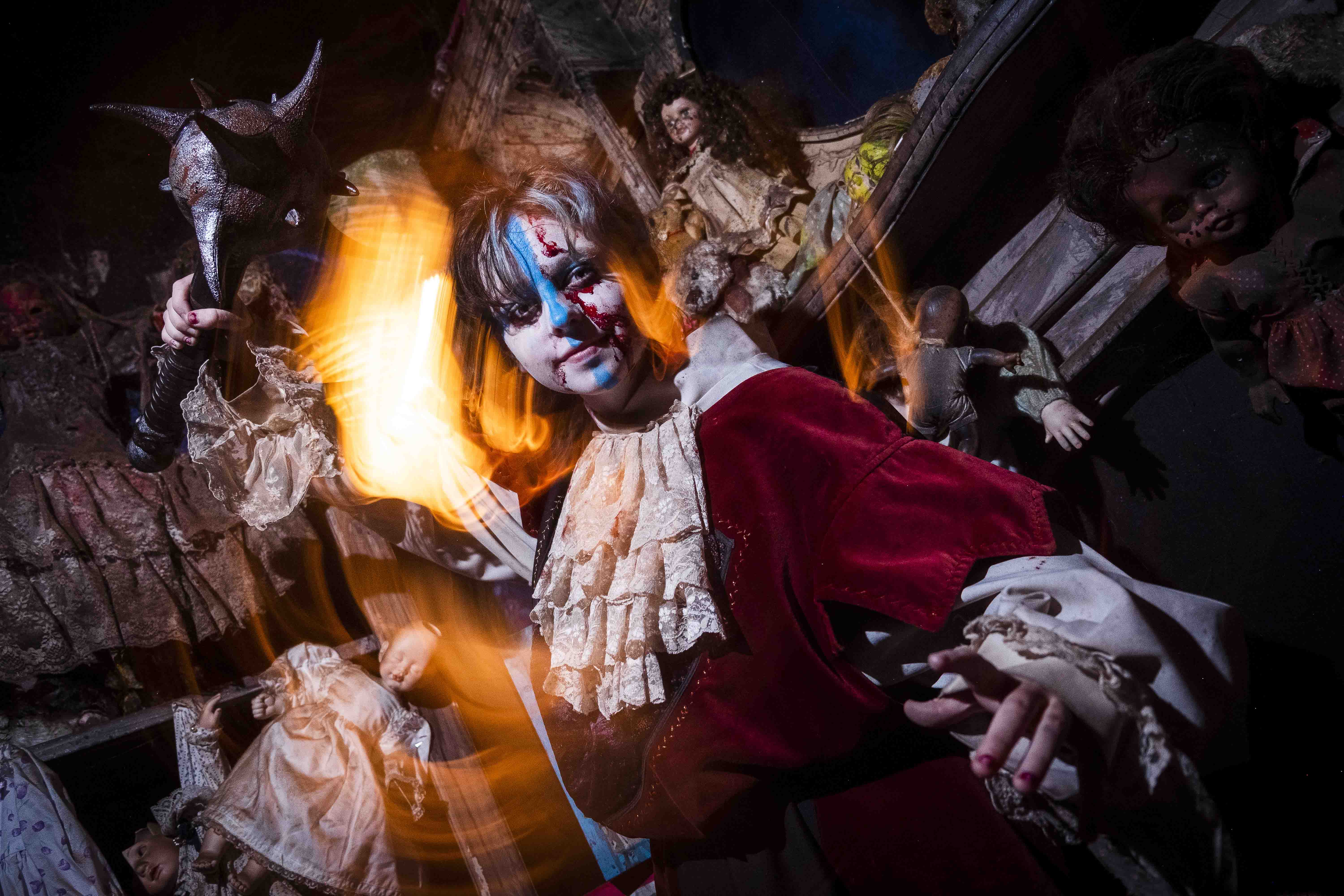 A person in a theatrical clown costume with dramatic face paint and a ruffled collar stands amidst a spooky, dimly-lit room filled with vintage dolls and eerie decorations, illuminated by warm, mysterious lighting, creating a haunting and captivating atmosphere.