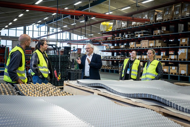 A group of people in safety vests inspecting shelves in a large warehouse. The atmosphere is busy and professional.