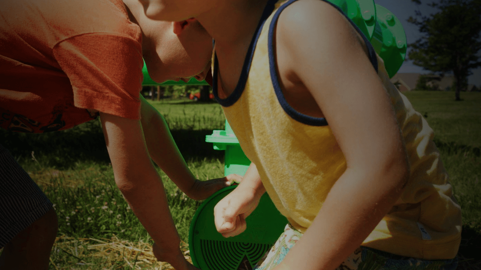 Two young children in park play with large green pipes that emerge from the grass