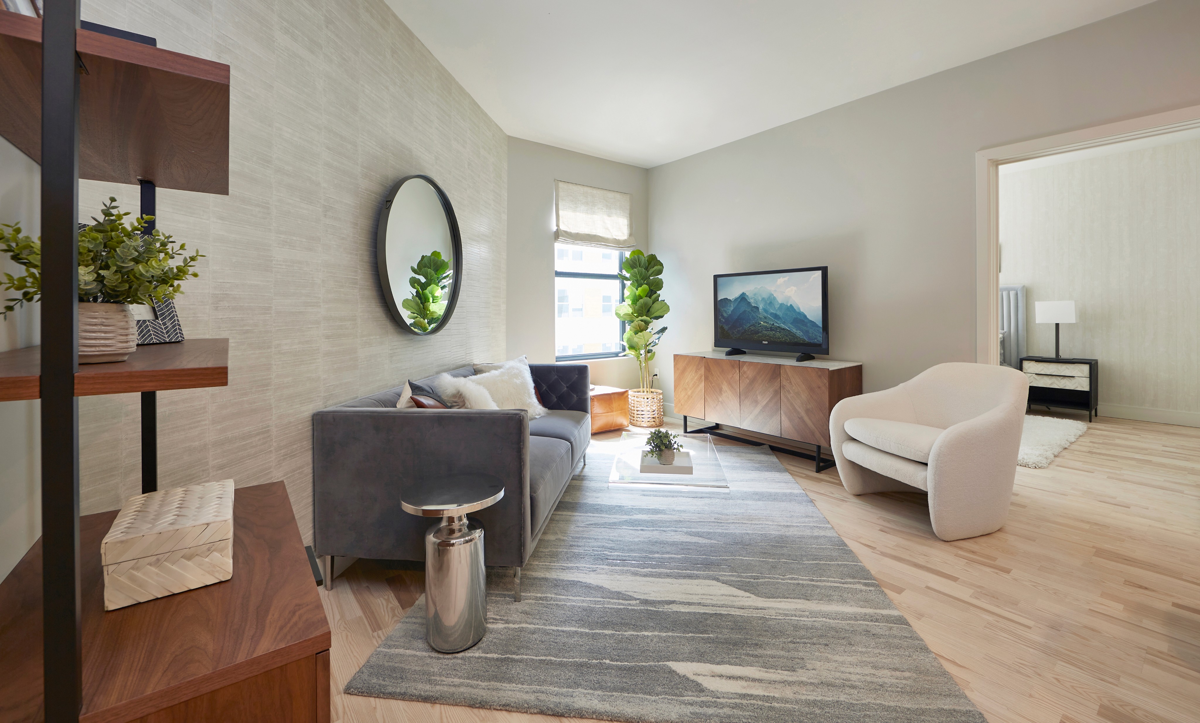 A stylish living room featuring a gray sofa, a round mirror, a TV, and modern decor, with natural light coming in.