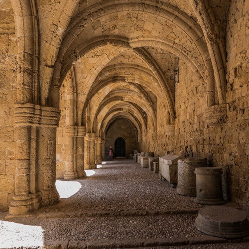 Stone archway corridor with vaulted ceiling, row of ancient tombs, and a distant dark door, lit by natural light from the left.