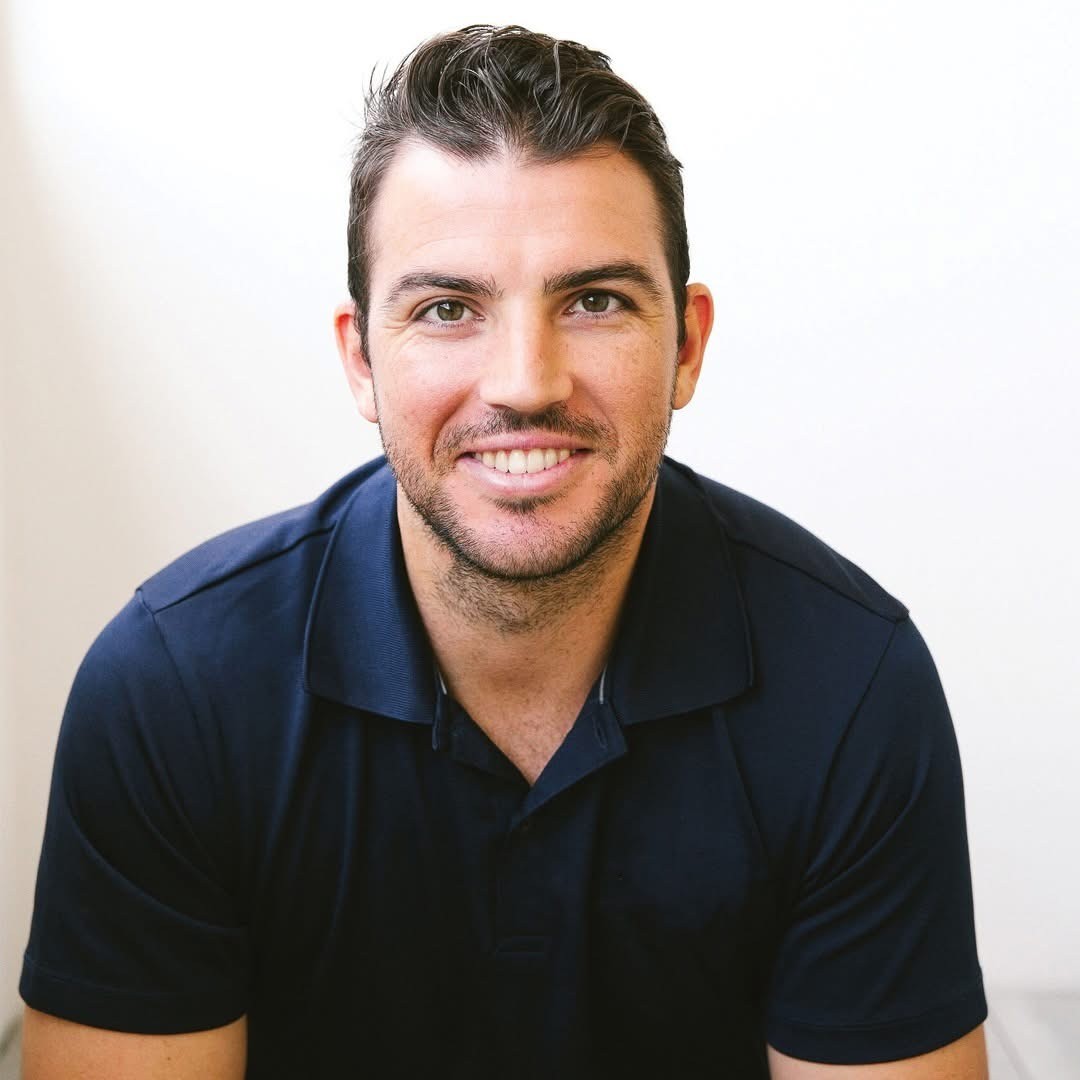 A person with curly hair and glasses smiles at the camera, seated in a cozy workspace with warm lighting.