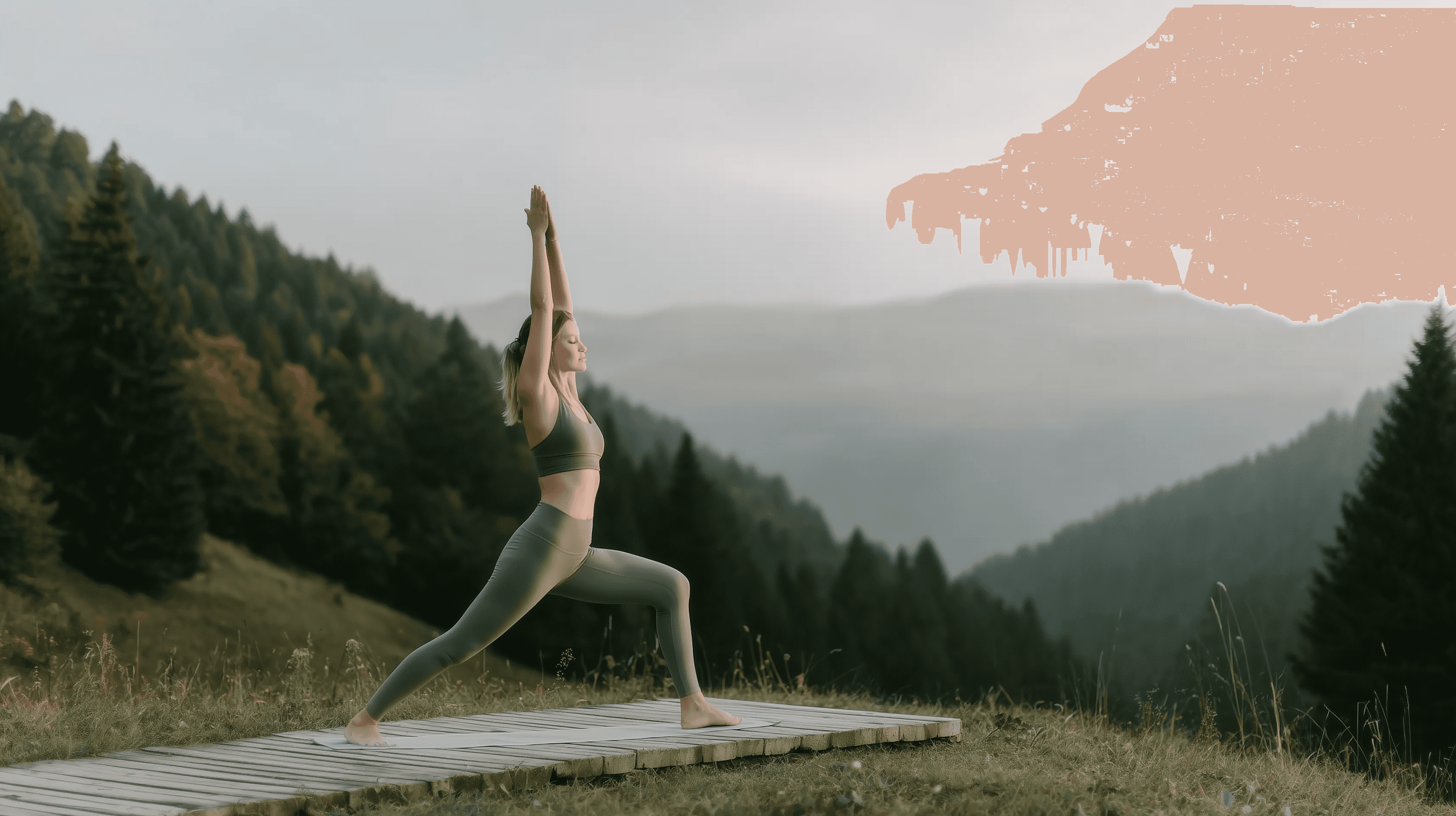 Woman practicing yoga warrior pose on a wooden platform surrounded by forested mountains in early morning light.