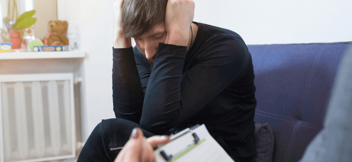A person dressed in black sits hunched forward on a blue sofa with their head in their hands in a posture of distress, while another person in the foreground holds a clipboard, suggesting a therapy or counselling session.