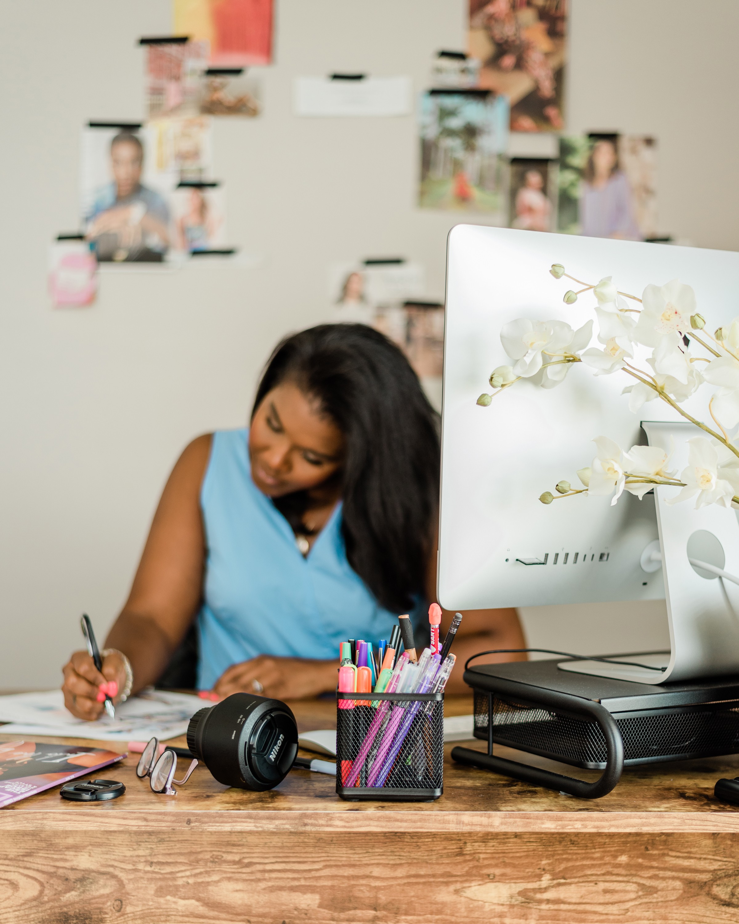 A person with curly hair and glasses smiles at the camera, seated in a cozy workspace with warm lighting.