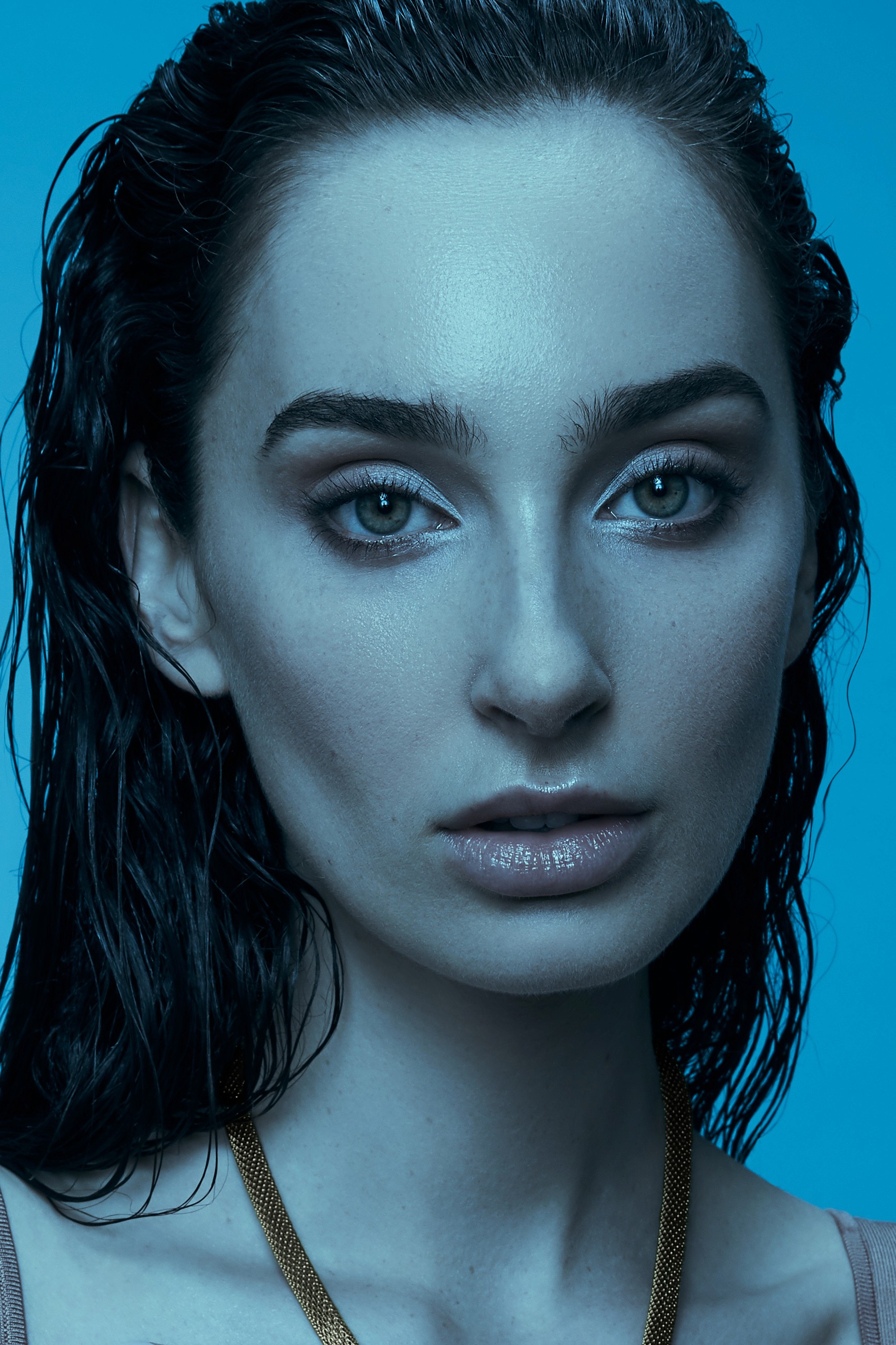 Beauty portrait in soft, blue light of a female model looking towards camera with damp hair, bare shoulers and a golden necklace that contrasts in tone with the rest of the image.
