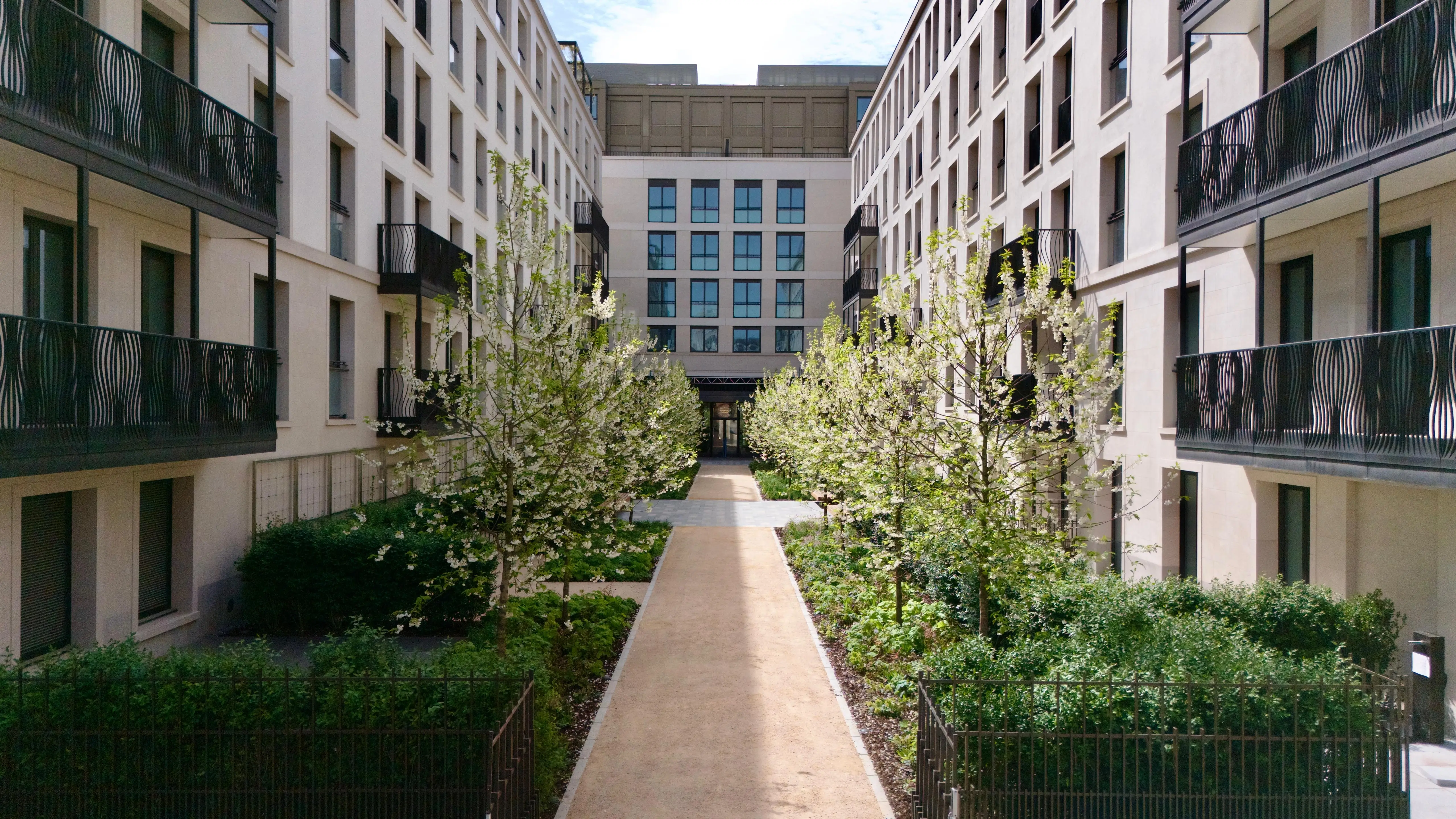 A paved pathway runs between two apartment buildings, lined with green bushes and trees under a clear sky.