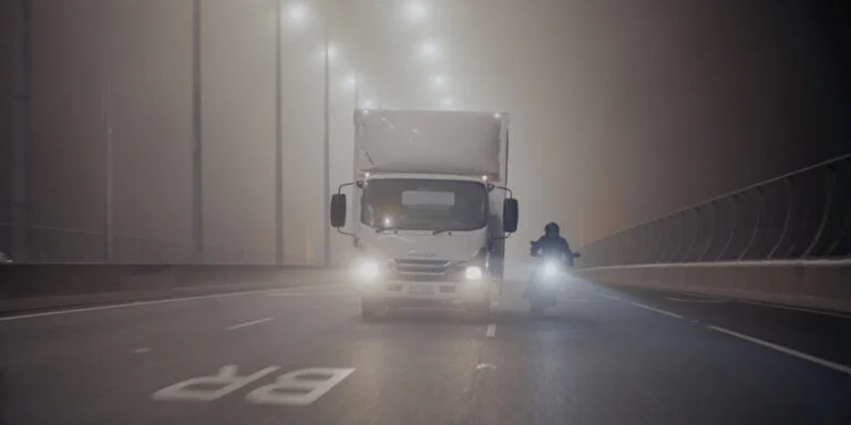 White Isuzu truck driving toward camera with headlights on along a foggy night road, with a motorbike alongside