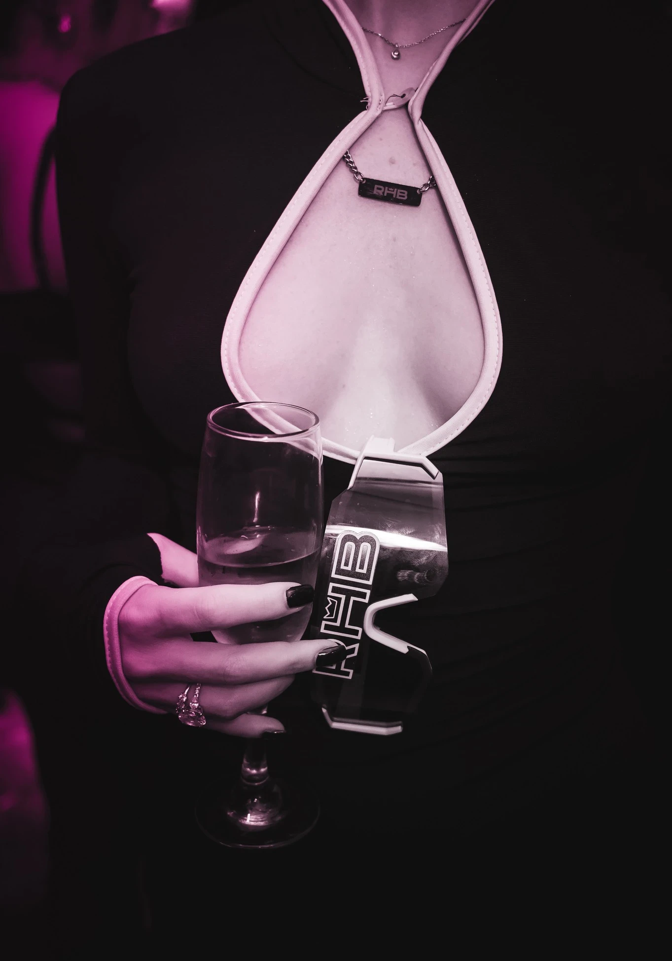 A close-up of a person at the RHB party holding a wine glass in one hand and a uniquely shaped, branded "RHB" cup in the other. They are wearing an "RHB" charm necklace.